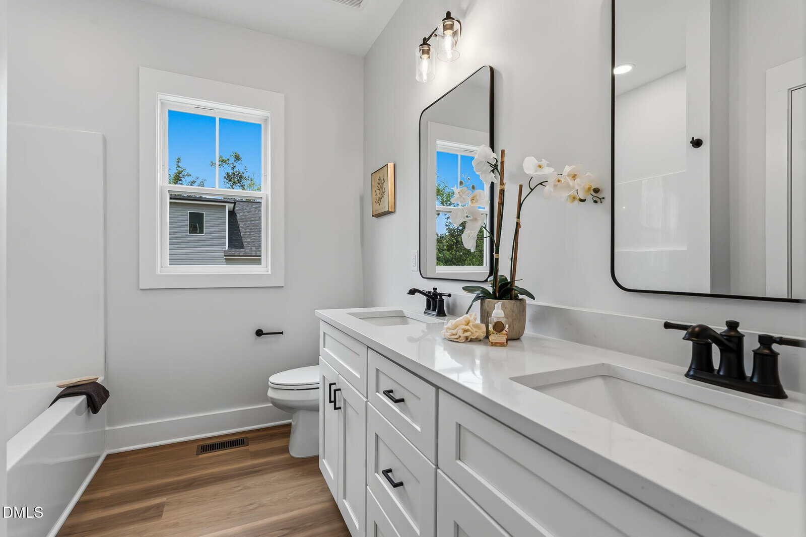 Modern bathroom with white cabinets, double sinks, black fixtures, a large mirror, a toilet, a window, a bathtub, and wood-style flooring. Orchids and shell decor are on the countertop. Walls and ceiling are white.