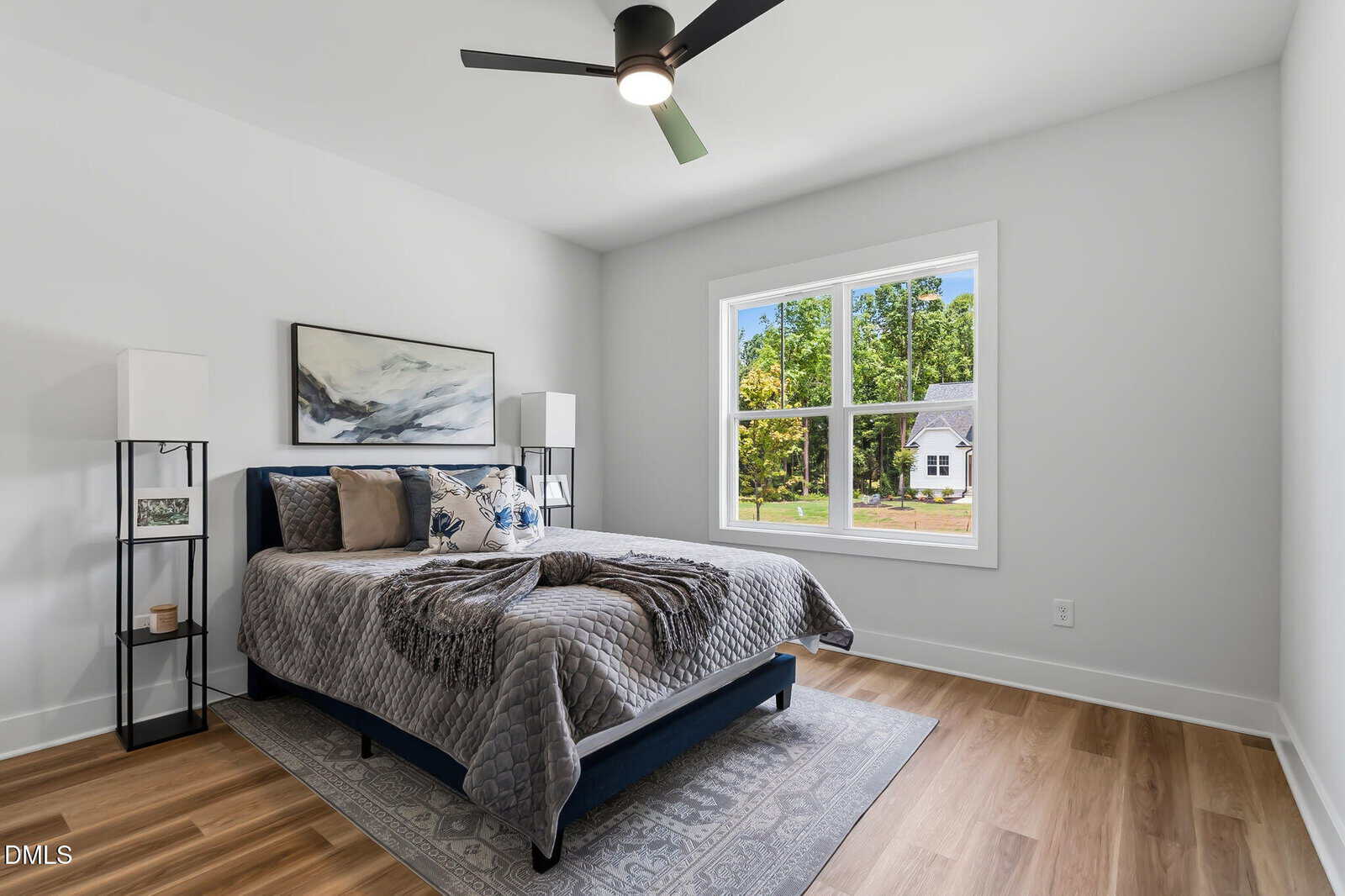 A modern bedroom with a bed, pillows, gray blanket, two tall lamps, abstract artwork above the bed, light wood floor, a small rug, and a large window showing a residential street with trees and houses.