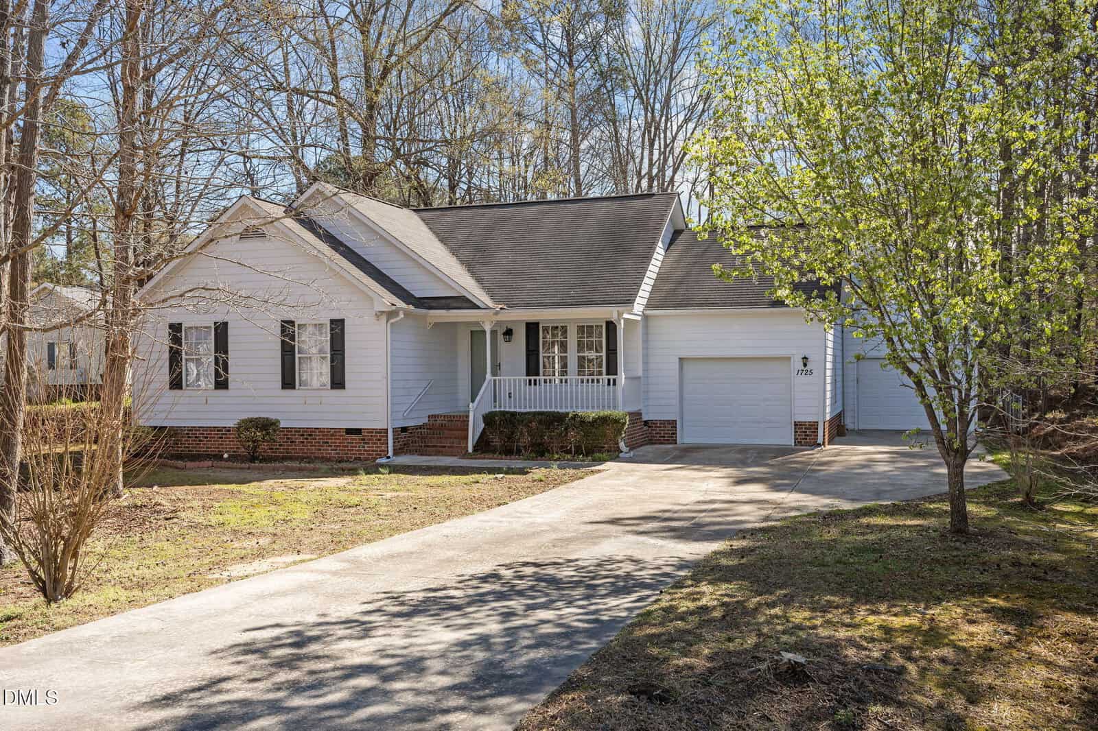 A single-story white house with a front porch, attached garage, and a sloped driveway. The yard has sparse grass, a few trees, and is bordered by woods in the background.