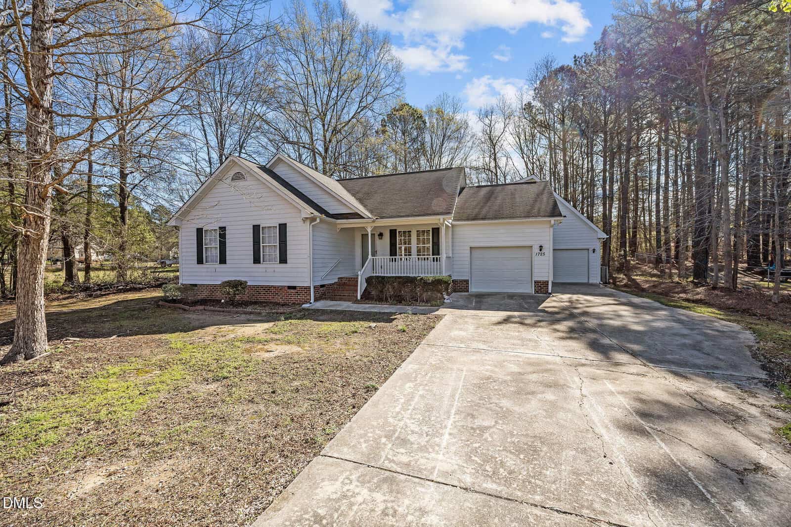 Single-story white house with a covered front porch, attached garage, and gabled roof, set among tall trees with a concrete driveway and a mostly bare front yard.
