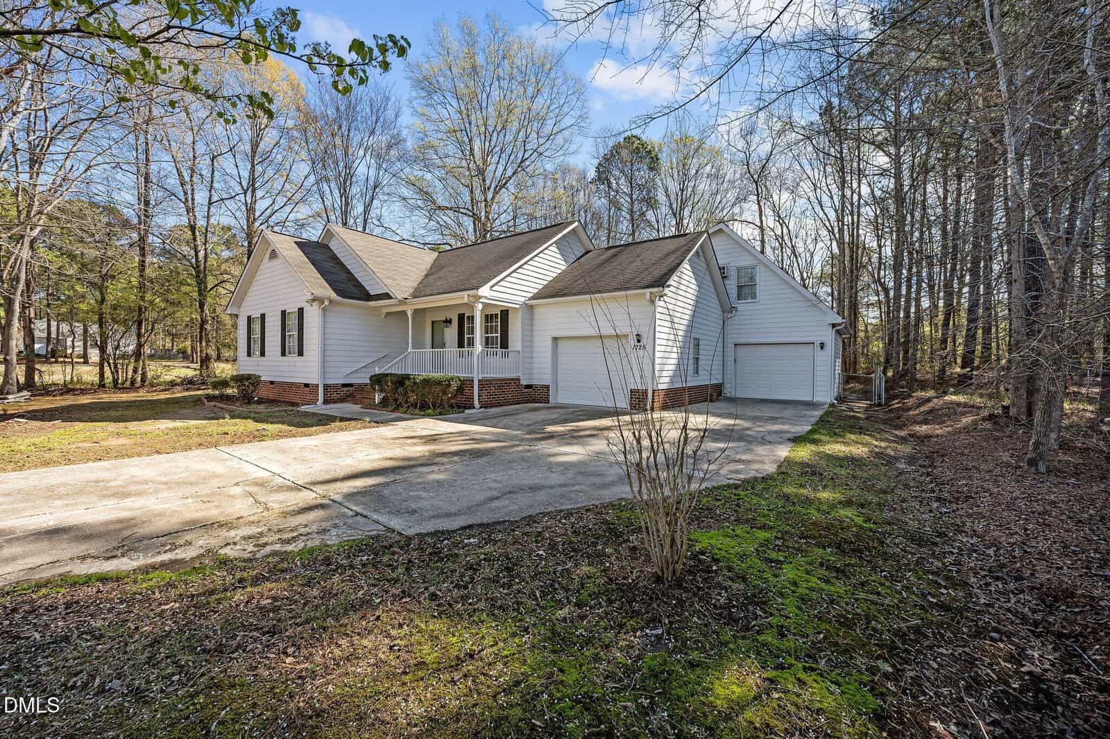 Single-story white house with a gabled roof, attached two-car garage, and brick foundation, surrounded by trees and a large driveway in a wooded area.