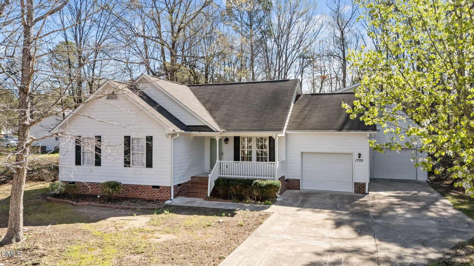 A single-story white house with a brick foundation, front porch, and attached garage. The driveway leads to the garage, and there are trees without leaves and some greenery around the property.