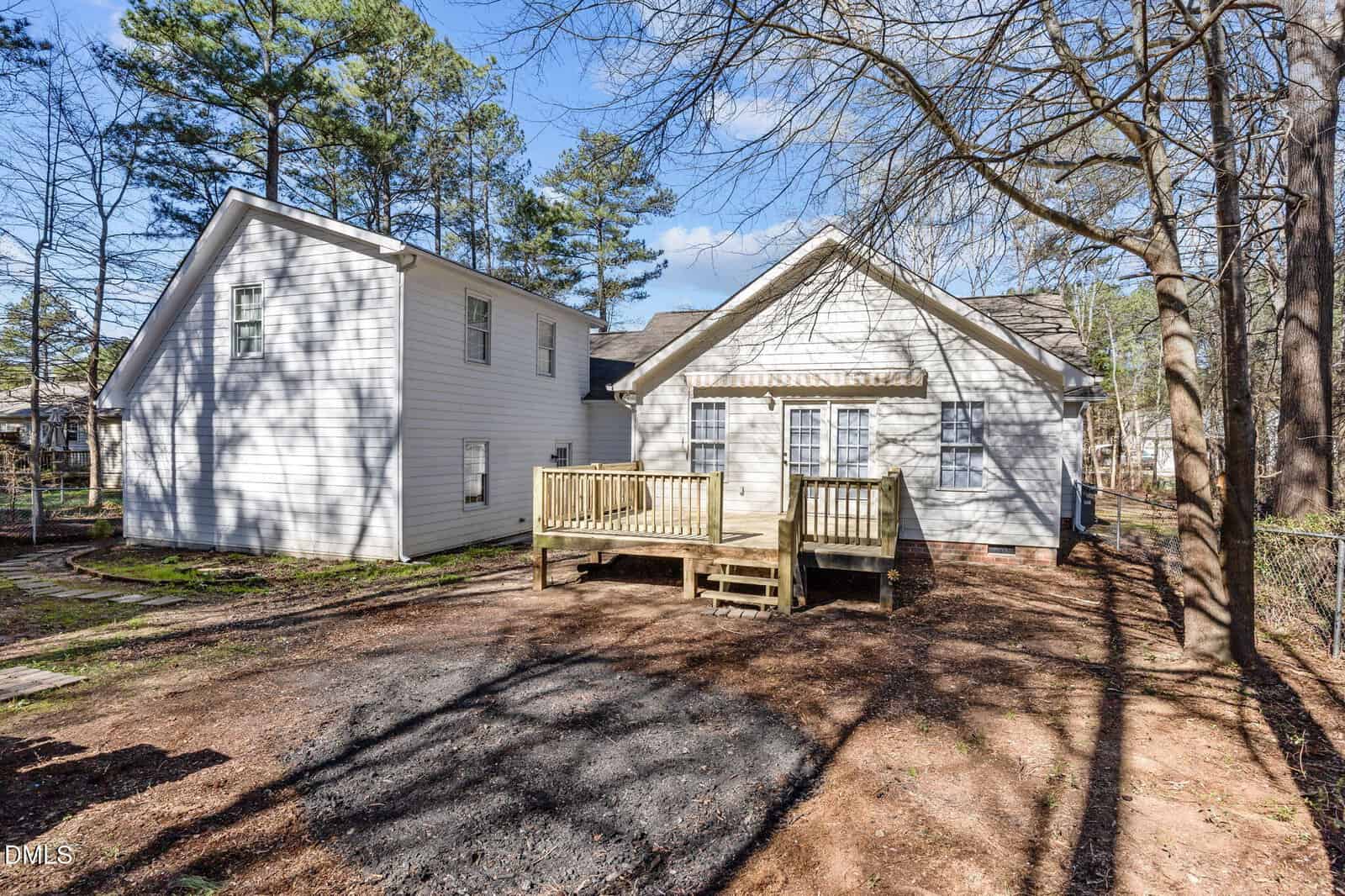 Single-story white house with two visible sections, a small wooden deck and stairs at the back, surrounded by trees and unpaved ground. Shadows from trees cover parts of the house and yard.