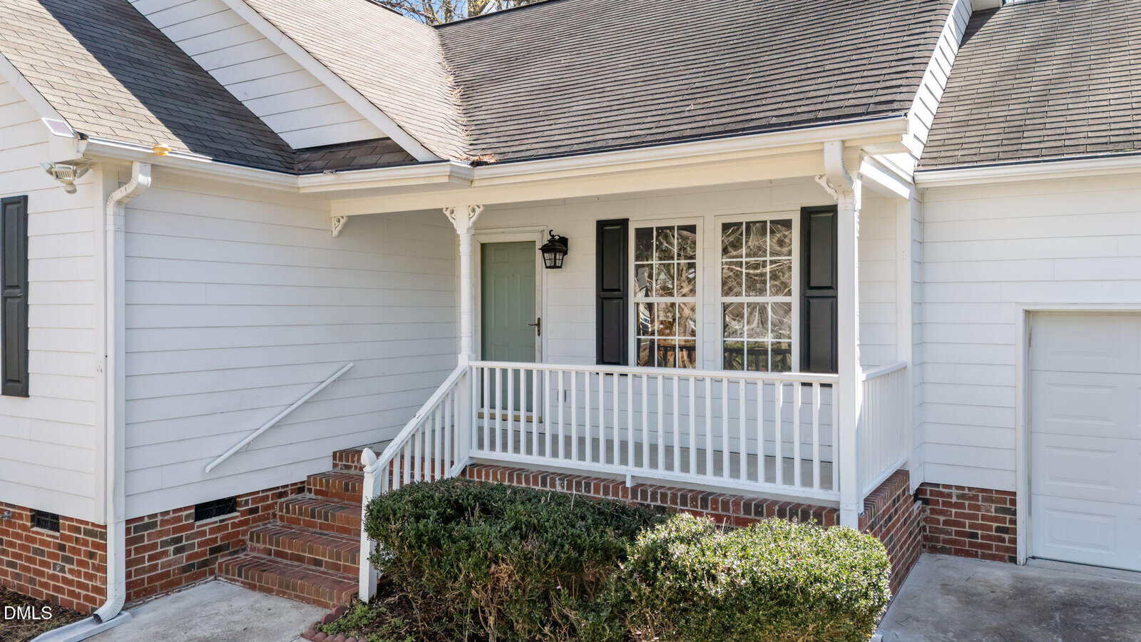 White house with black shutters, a porch with white railing, brick steps, and a small bush in front. The entry door is next to a window with grid panes. Part of the garage door is visible on the right.