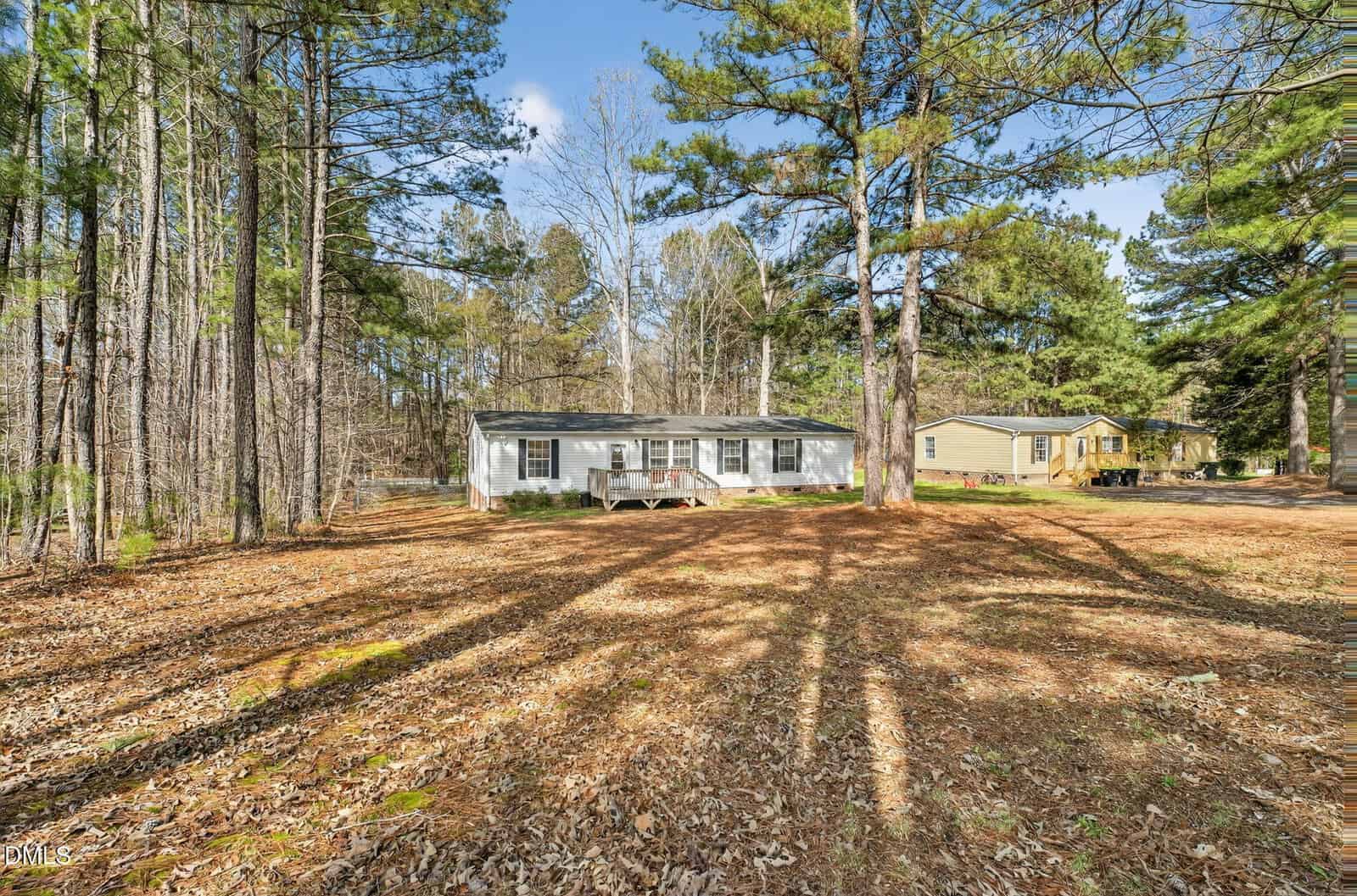 A single-story white house with a small front porch is surrounded by tall pine trees and fallen leaves. Another house and a yellow vehicle are visible in the background on a sunny day.
