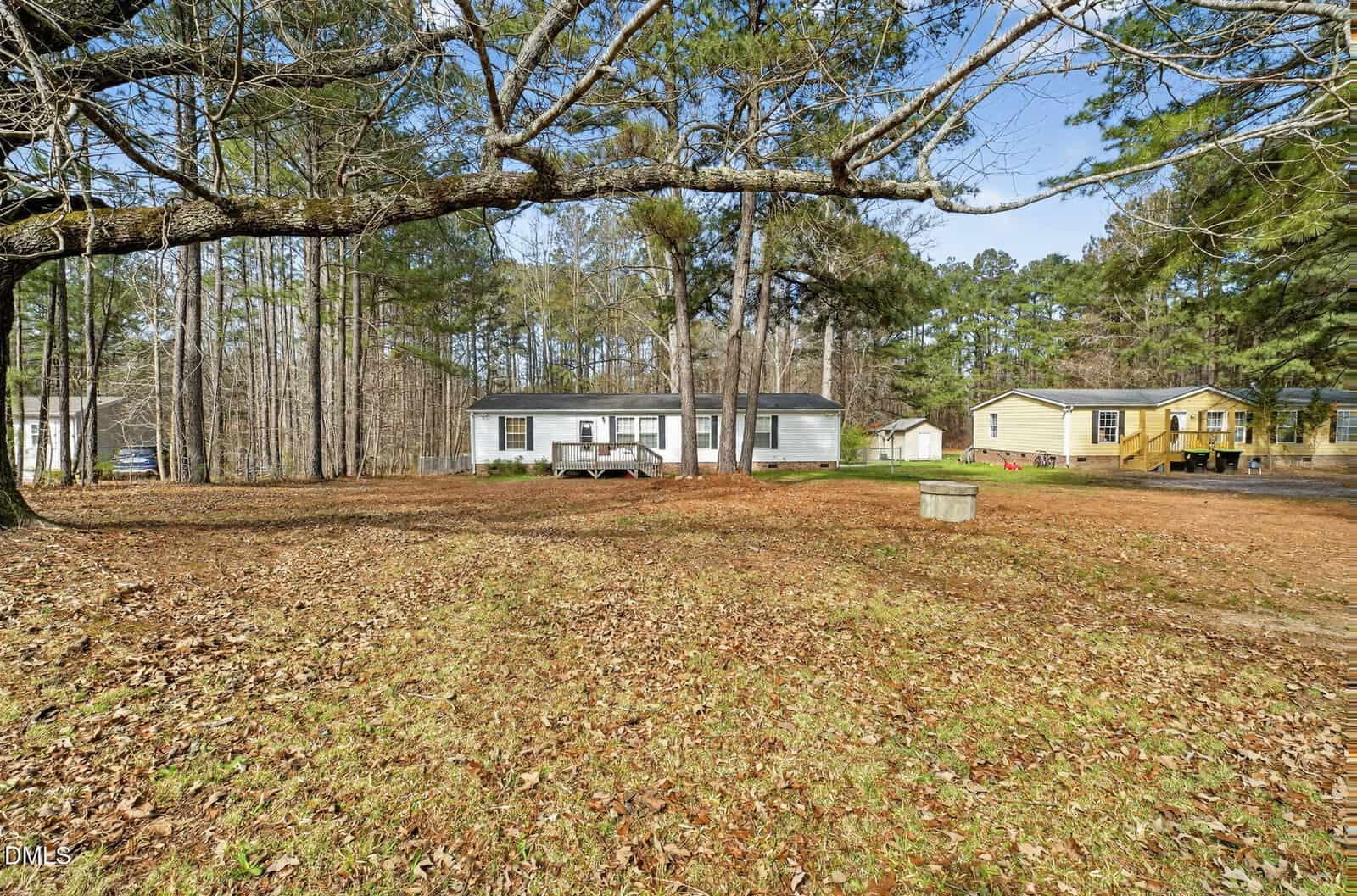 A one-story white house with a small front porch sits in a large, grassy yard with scattered fallen leaves. Trees surround the property, and a yellow house is visible to the right. The sky is clear.