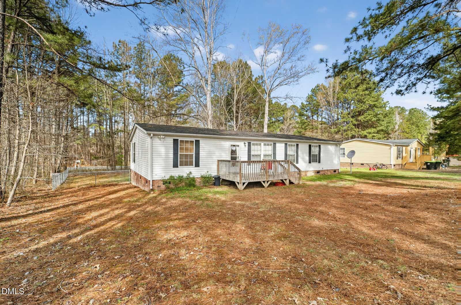 A single-story manufactured home with light-colored siding and a small wooden front porch is set on a grassy lot with tall pine trees. Another similar house is visible in the background. The sky is clear and sunny.