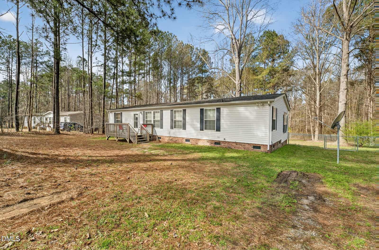 A white, single-story manufactured home with black shutters sits on a grassy lot surrounded by tall trees. A small wooden entry deck leads to the front door. The sky is mostly clear with some clouds.