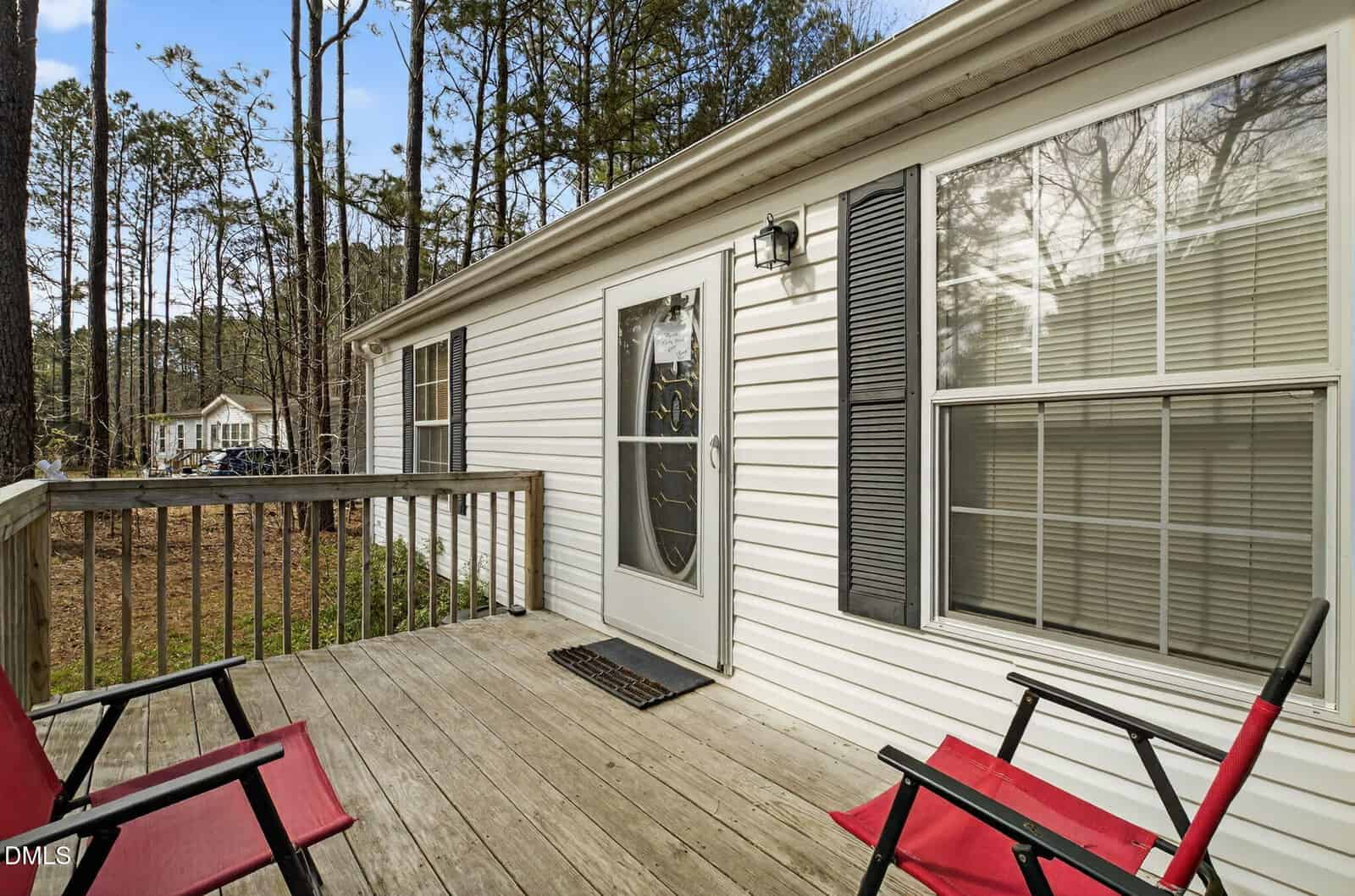 A small wooden deck with two red chairs faces a white house with horizontal siding, a glass door, a large window with blinds, black shutters, and a wall-mounted light. Trees and another house are visible in the background.