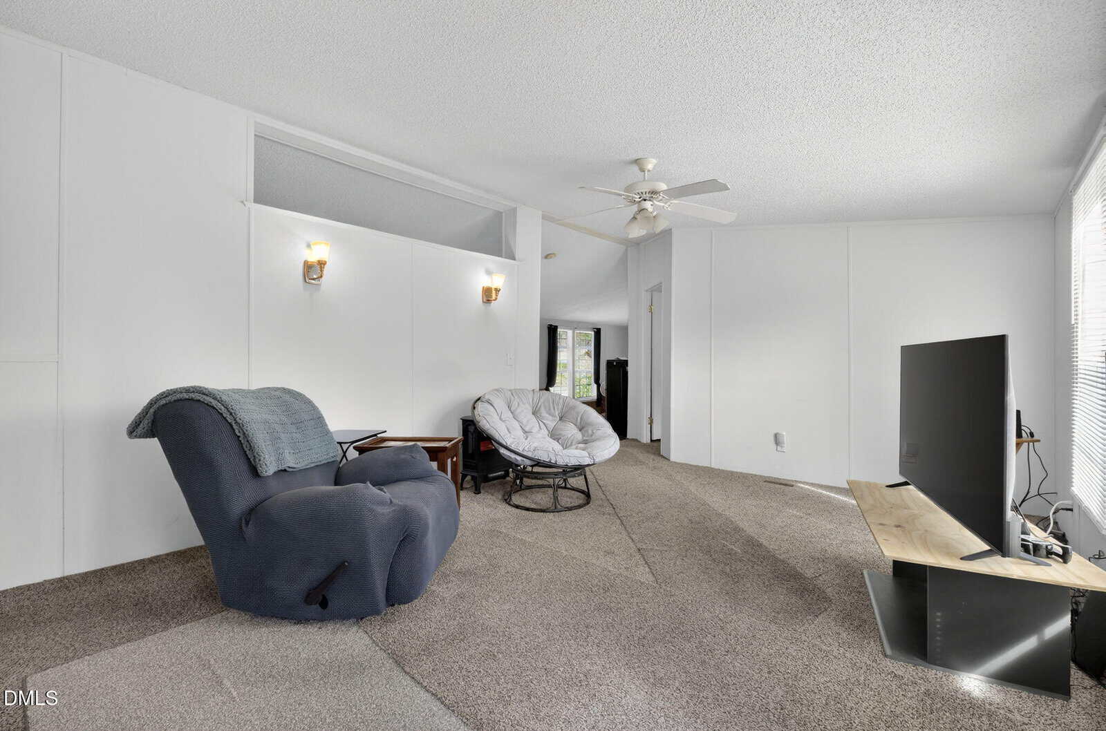 A living room with beige carpet, white walls, a blue recliner, a round cushioned chair, a TV on a wooden stand, and a ceiling fan. Two wall sconces provide light. A doorway is visible in the back.