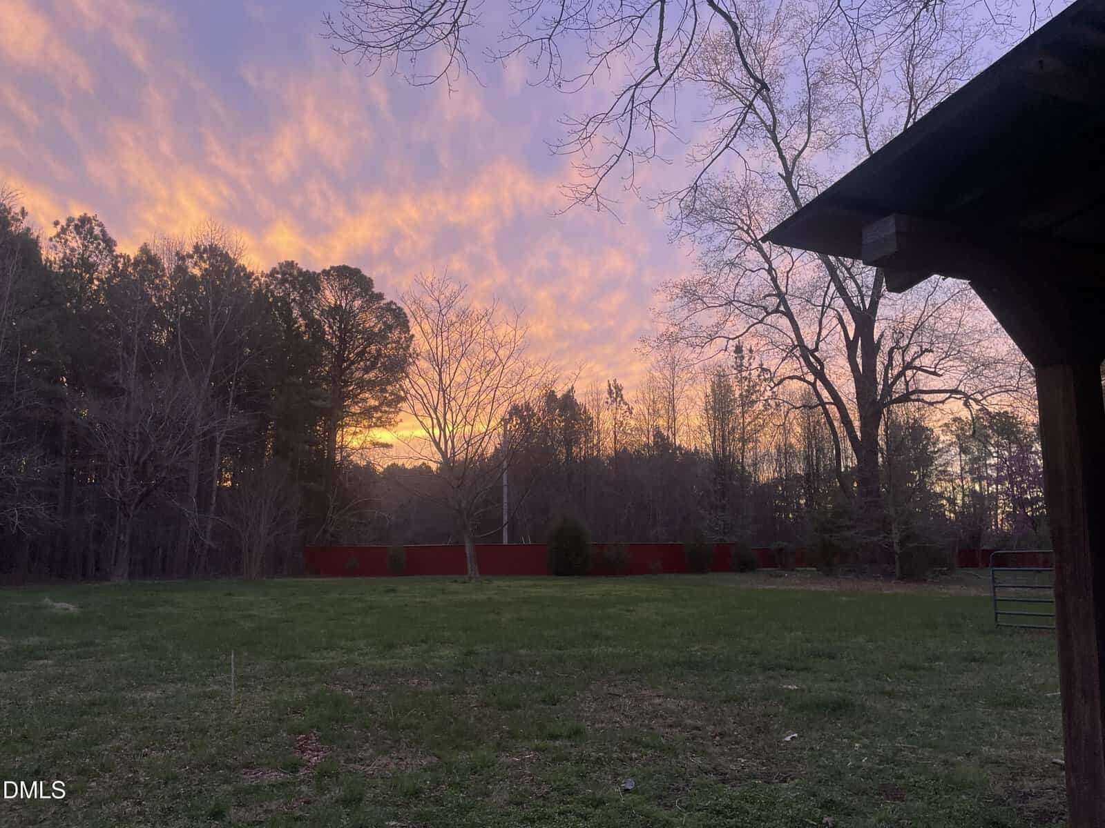 A grassy backyard with scattered trees and a wooden structure on the right, bordered by a red fence, under a colorful sunset sky with pink, orange, and purple clouds.