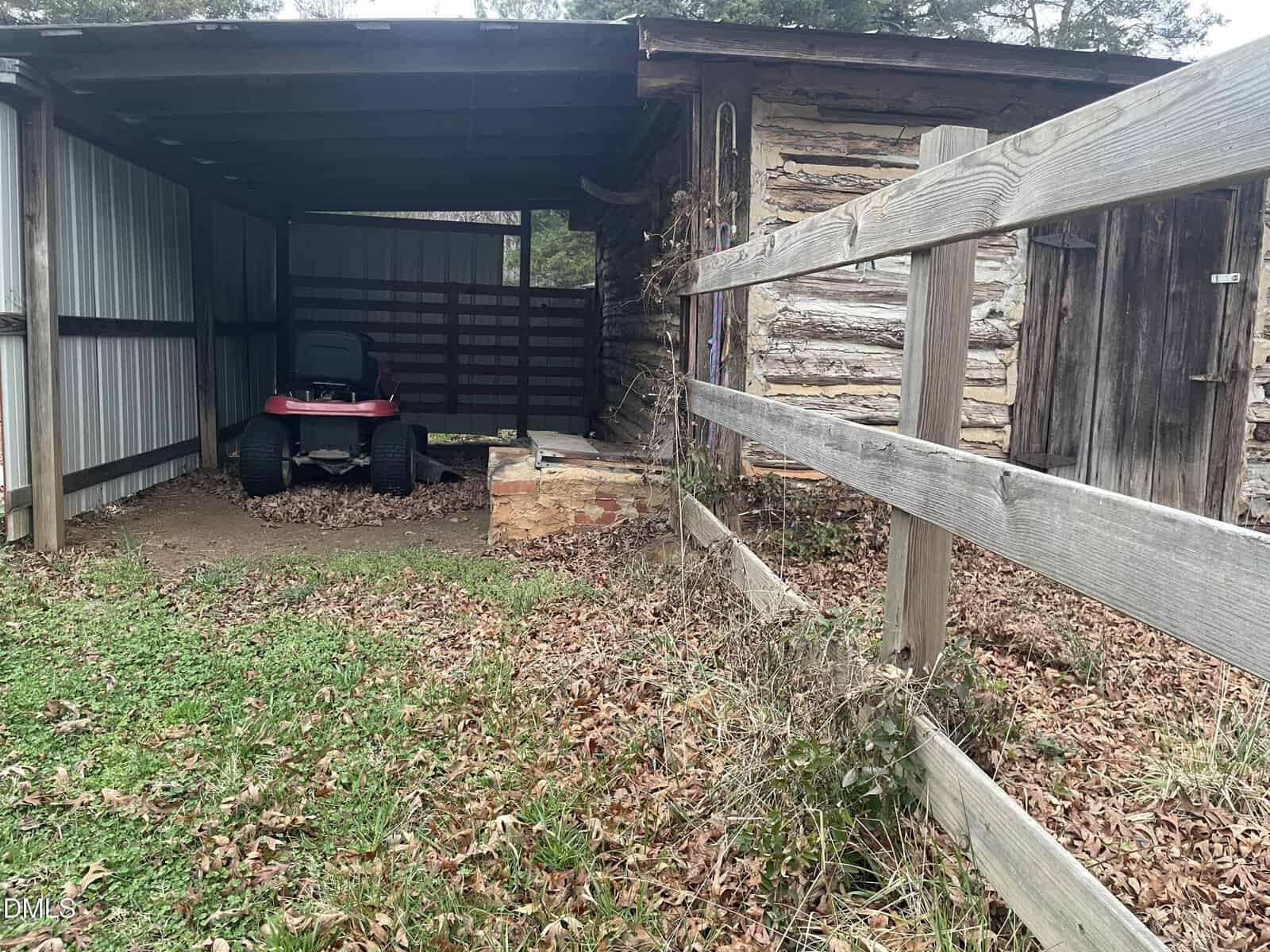A red riding lawn mower is parked in a covered shed next to a wooden fence and a log cabin. Fallen leaves are scattered on the ground, and some grass is visible in the foreground.