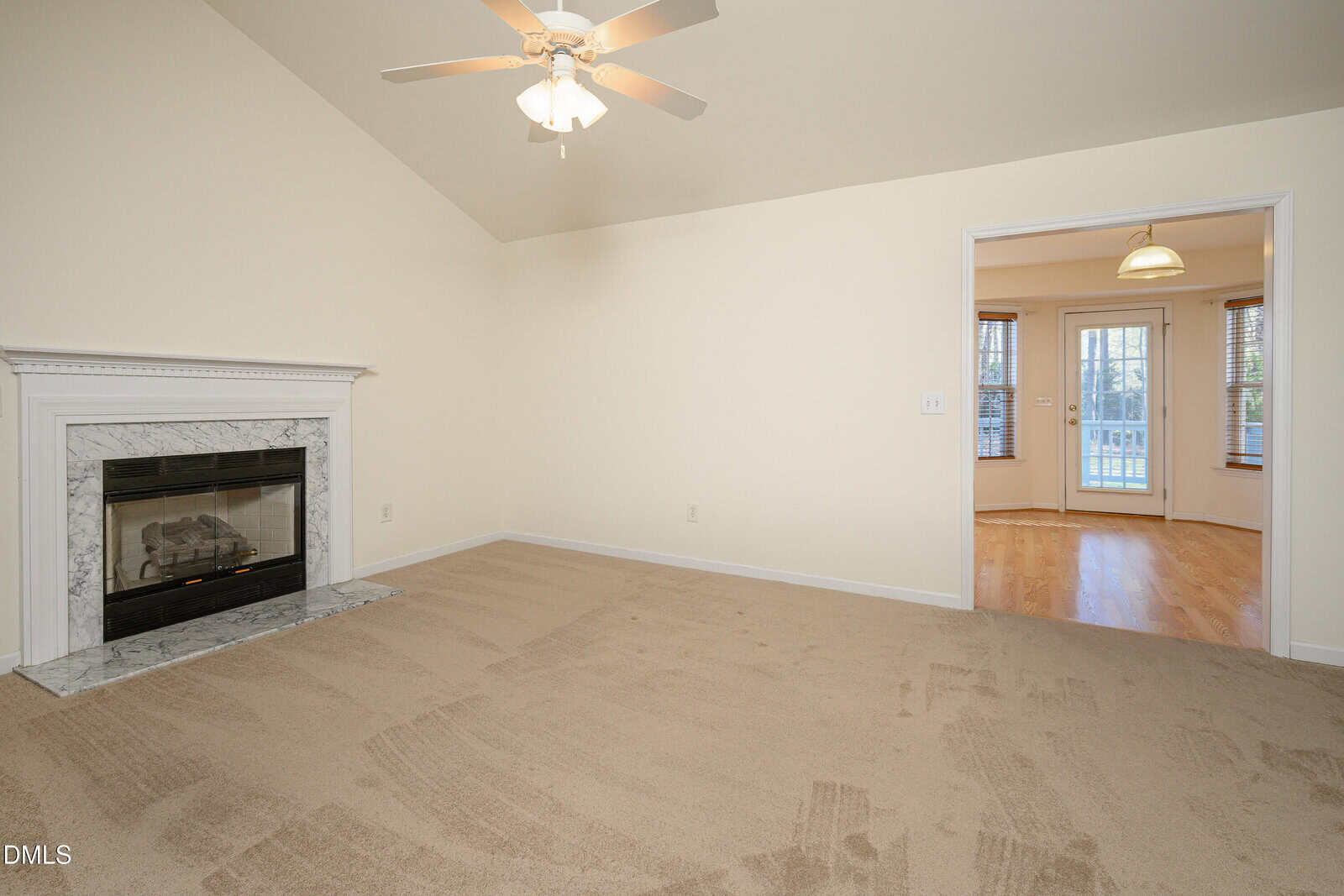 A carpeted living room with a marble fireplace on the left wall, a ceiling fan with lights, and an open doorway leading to a sunlit room with hardwood floors and large windows.