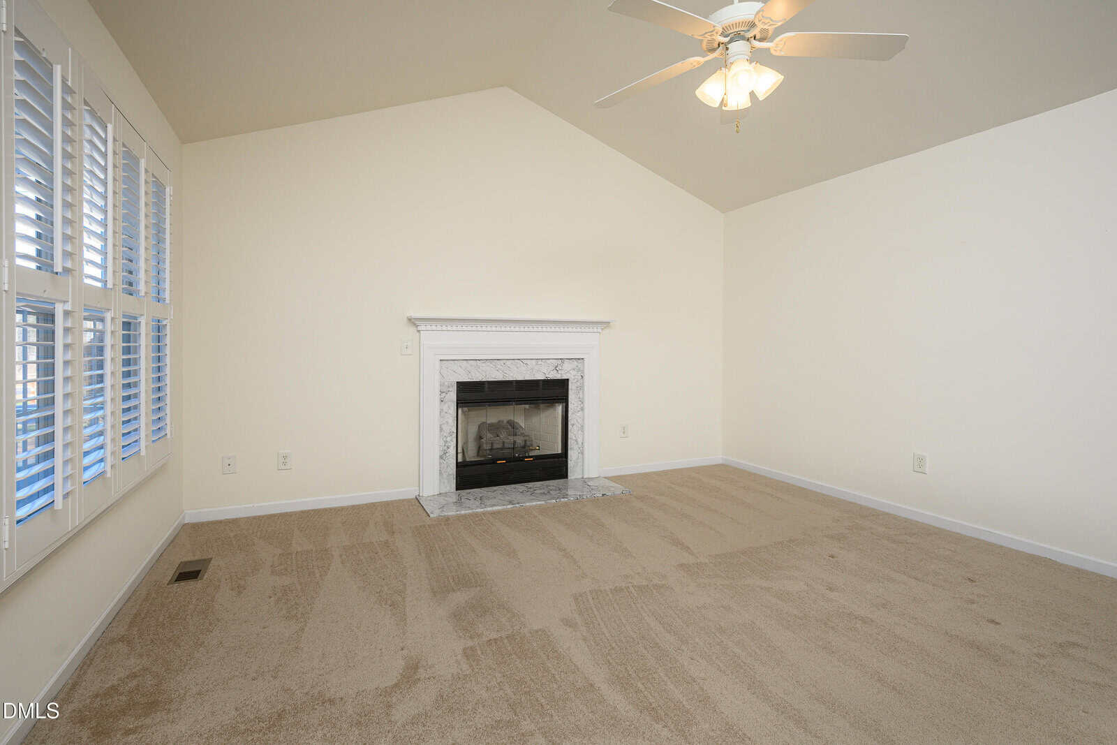 A vacant room with beige carpet, cream-colored walls, a white ceiling fan, white plantation shutters on the left wall, and a white fireplace with a marble surround centered on the far wall.