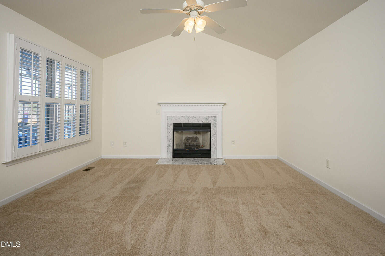 A carpeted living room with a white ceiling fan, a fireplace with a white mantel, and shuttered windows on the left wall. Walls are light beige, and the room appears empty.