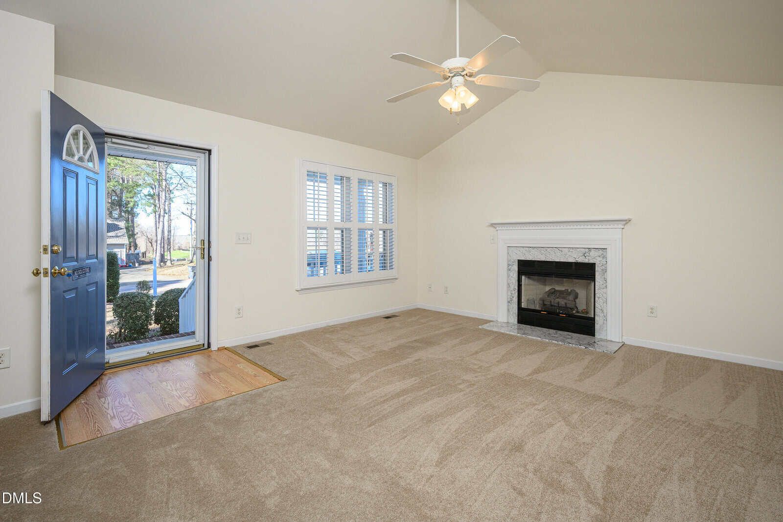 A carpeted living room with an open blue front door, a large window with white shutters, a white ceiling fan, and a white fireplace with a marble surround. The walls are painted beige.