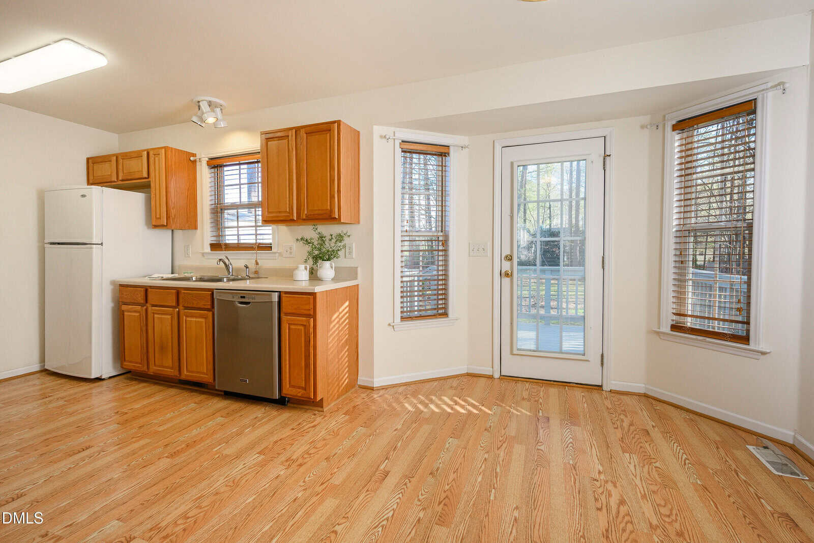 Bright kitchen with wooden cabinets, stainless steel dishwasher, white refrigerator, sink, and large windows. A glass door leads to an outdoor area. Hardwood floors and white walls complete the room.