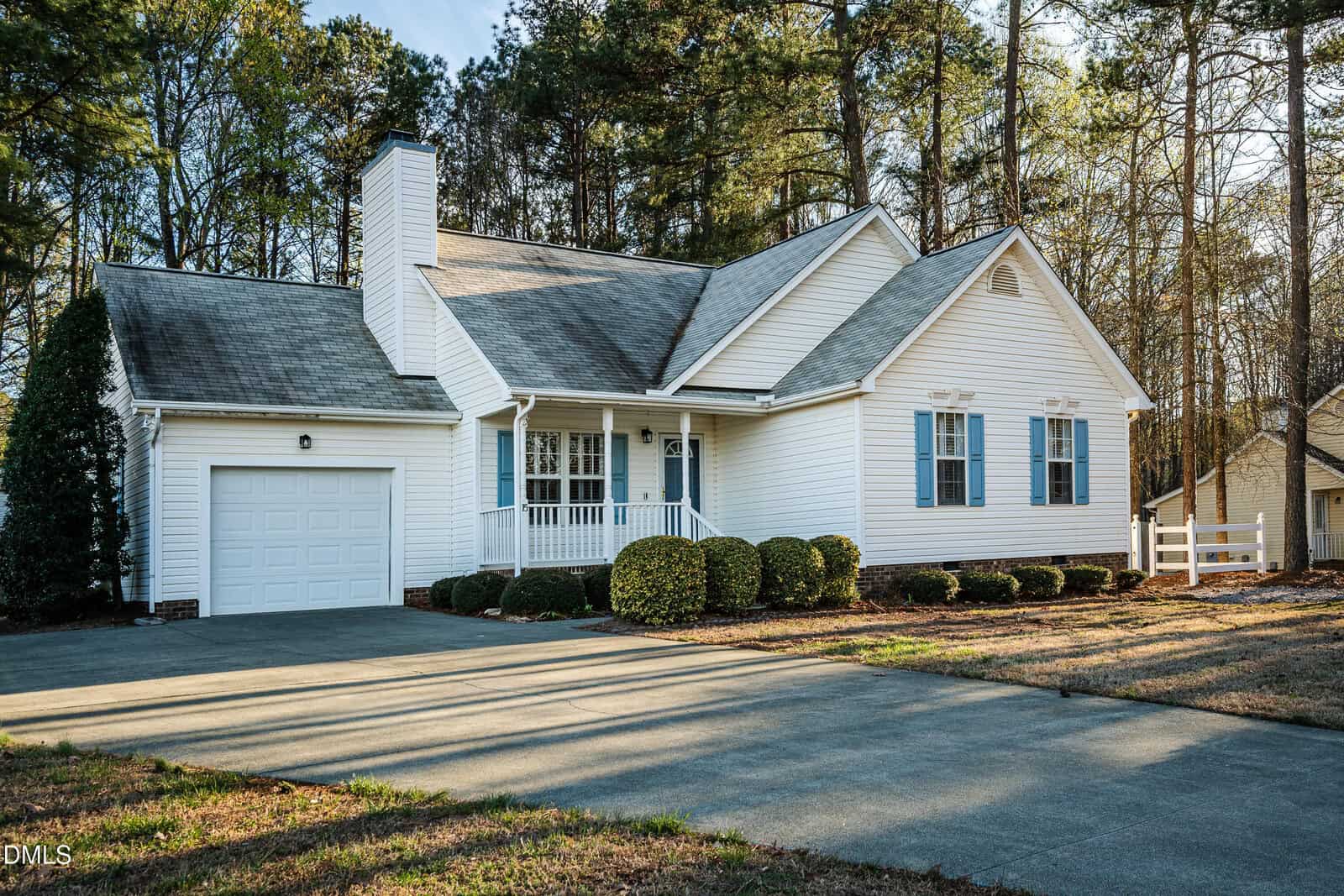 Single-story white house with blue shutters, attached garage, front porch, and driveway. The yard has manicured bushes and is surrounded by tall trees. It is a sunny day with clear skies.