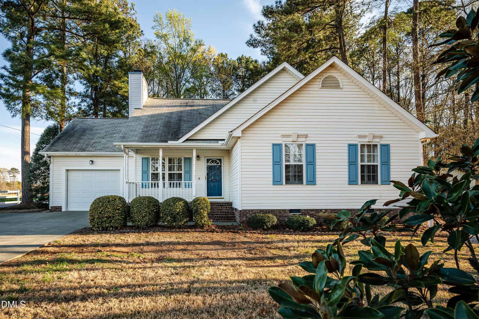 A single-story white house with blue shutters and a front porch, attached garage on the left, surrounded by bushes and tall trees under a clear sky.
