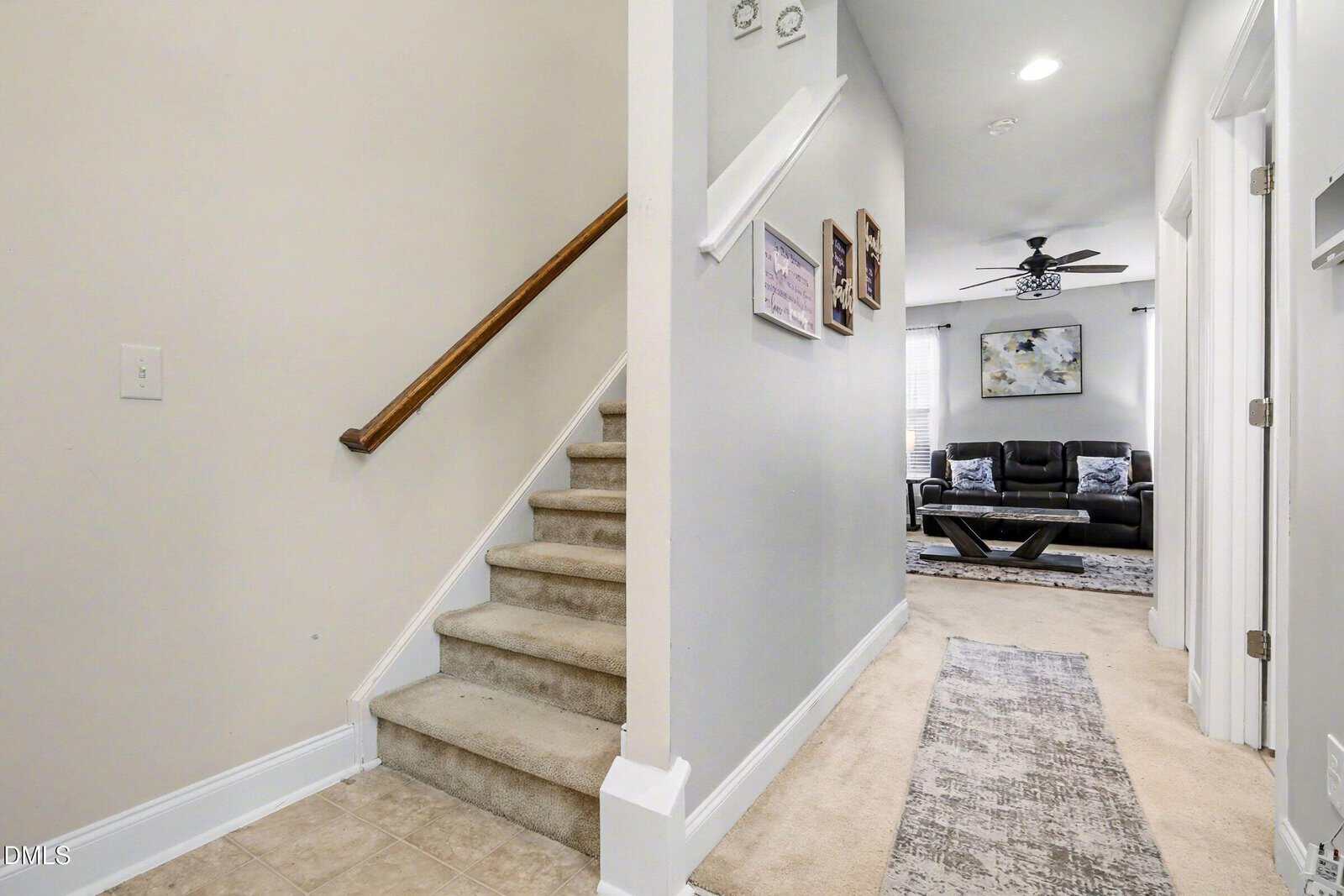 Carpeted stairs with a wooden handrail lead upward beside a hallway. The hall, decorated with framed pictures, opens into a living room with a dark sofa, coffee table, wall art, ceiling fan, and windows with blinds.