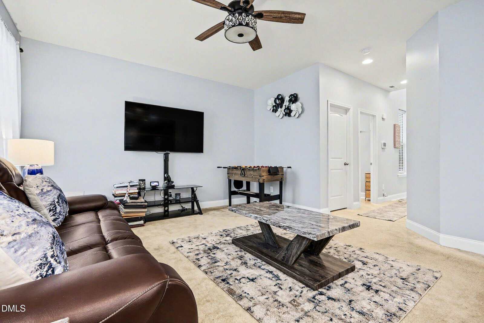 A living room with a brown leather sofa, patterned pillows, a wooden coffee table, a TV mounted on the wall, a foosball table, and beige carpet. A ceiling fan and two rugs are also visible in the room.