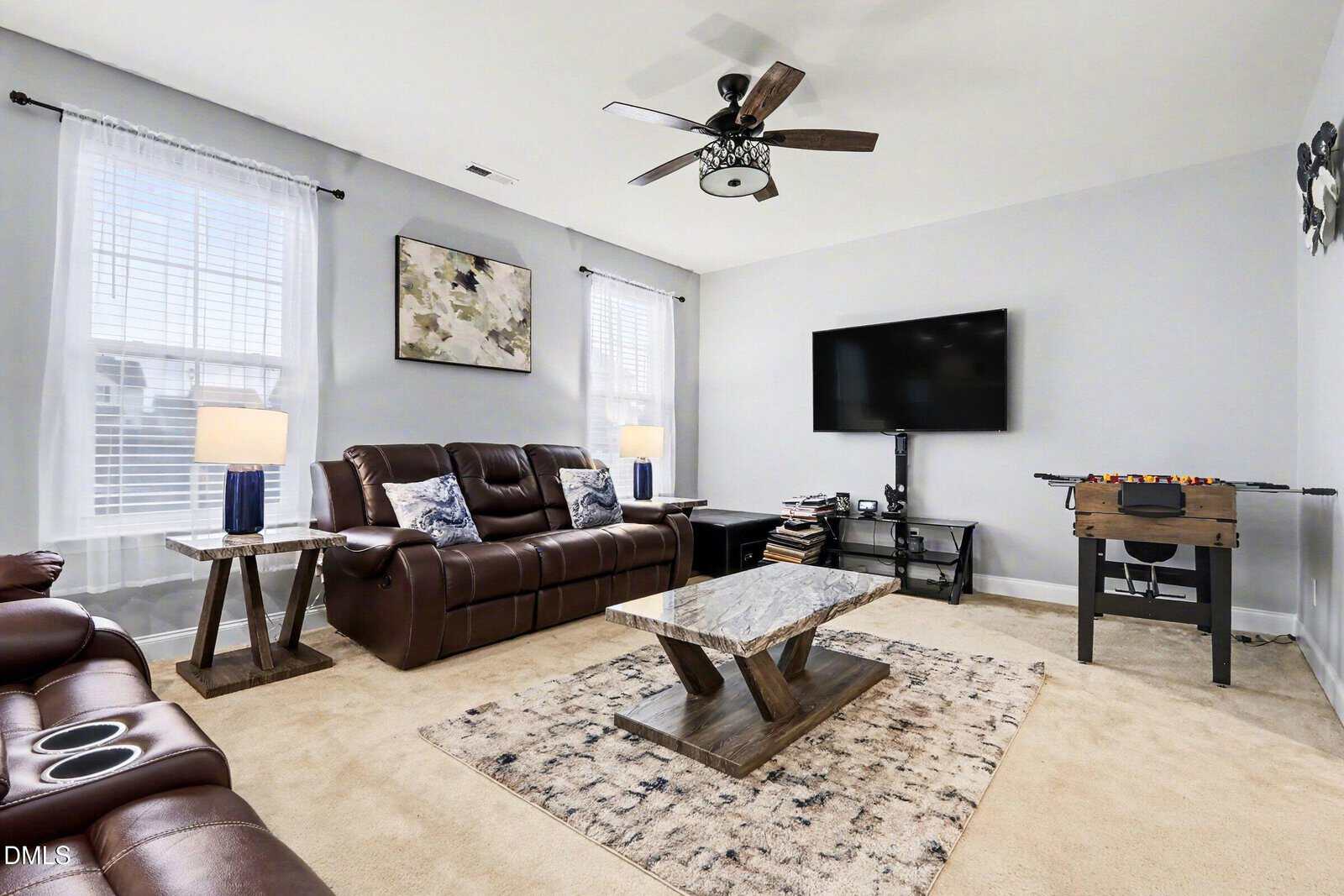 A living room with brown leather sofas, a marble coffee table, a rug, a TV on a stand, end tables with lamps, a foosball table, and wall art. The carpet is beige, and the walls are painted light gray.