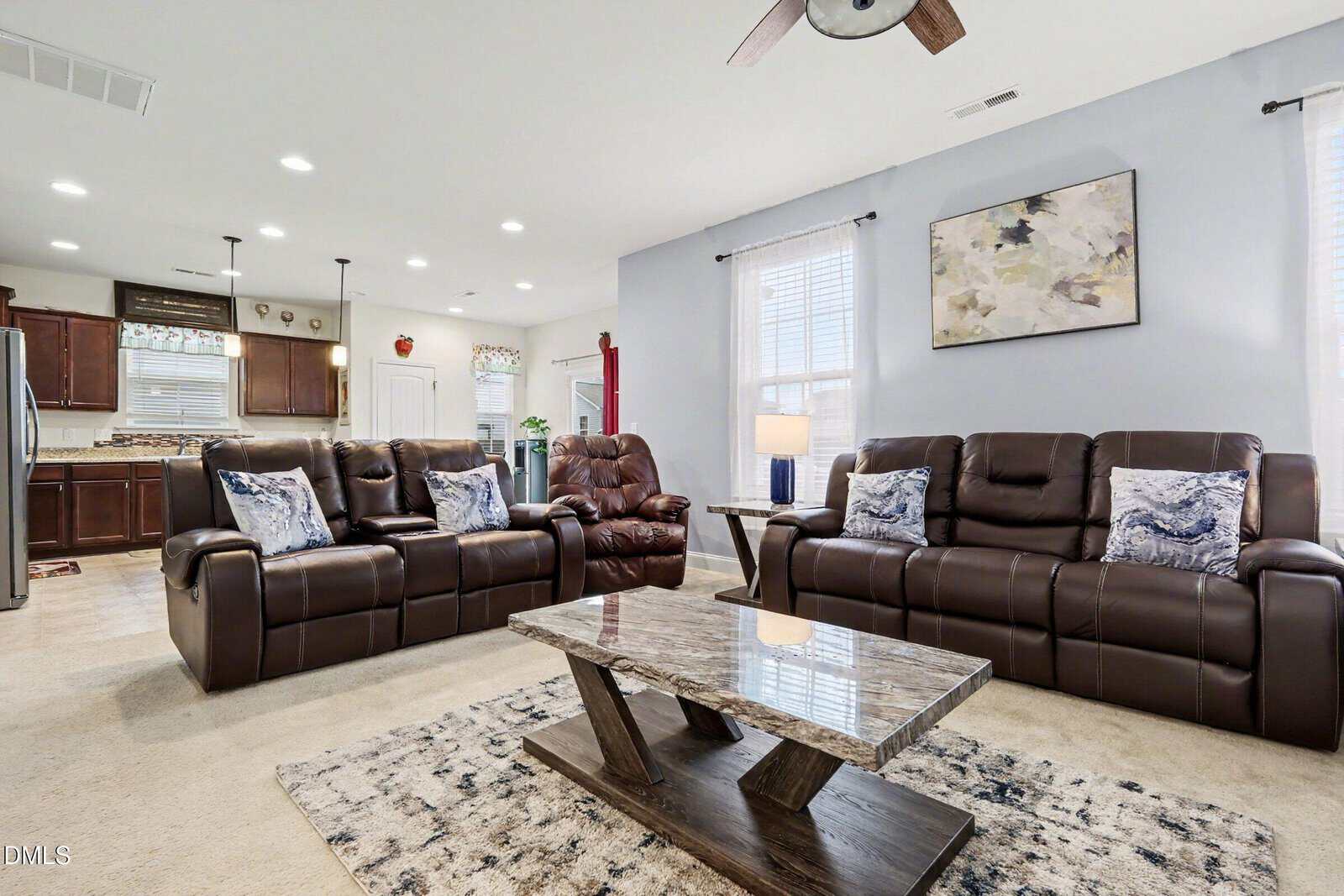 Modern living room with two brown leather sofas, a matching recliner, a marble coffee table, and a neutral rug. The space is open to a kitchen with dark cabinets and stainless steel appliances in the background.
