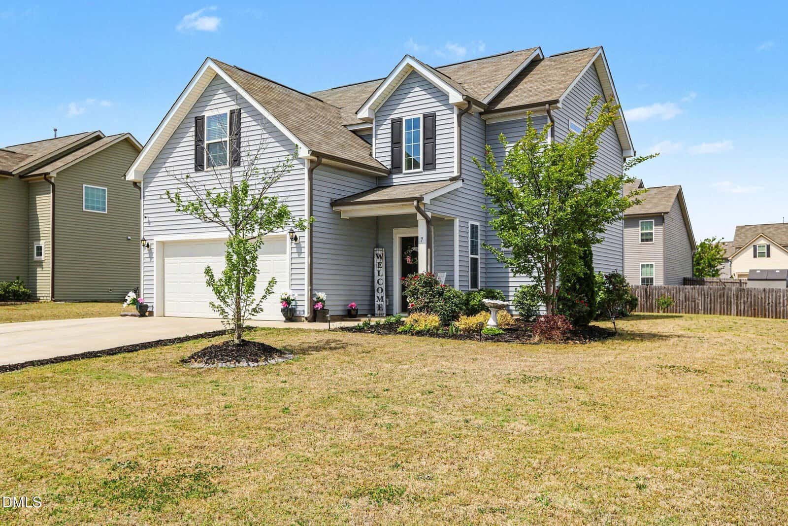 Two-story suburban house with gray siding, attached double garage, and a covered front porch. The front yard has a few small trees and shrubs, with neatly trimmed grass and clear blue sky above.
