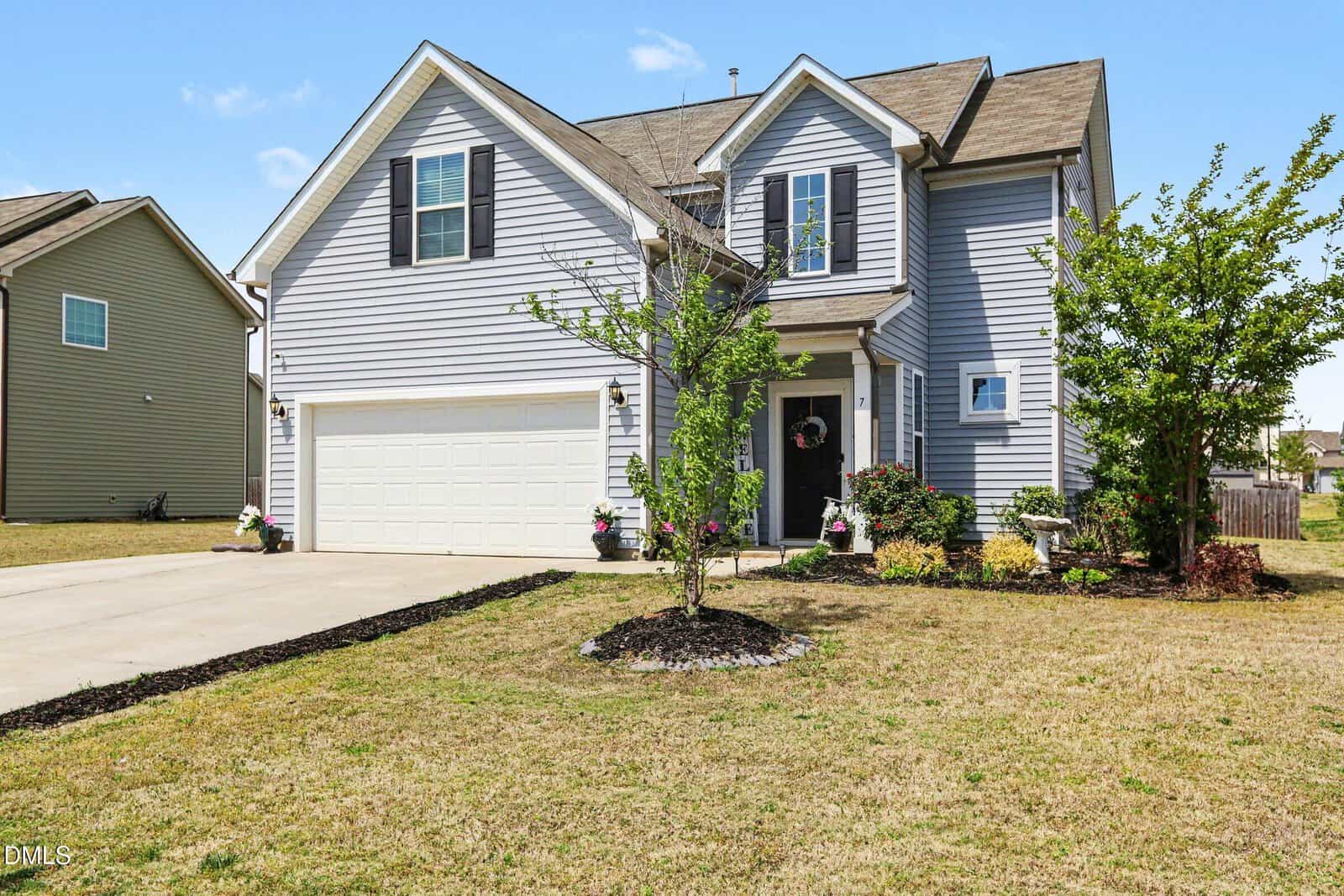Two-story gray house with white trim, black shutters, and a two-car garage. The front yard has a small tree, flower beds, and a concrete driveway. The sky is clear and sunny.