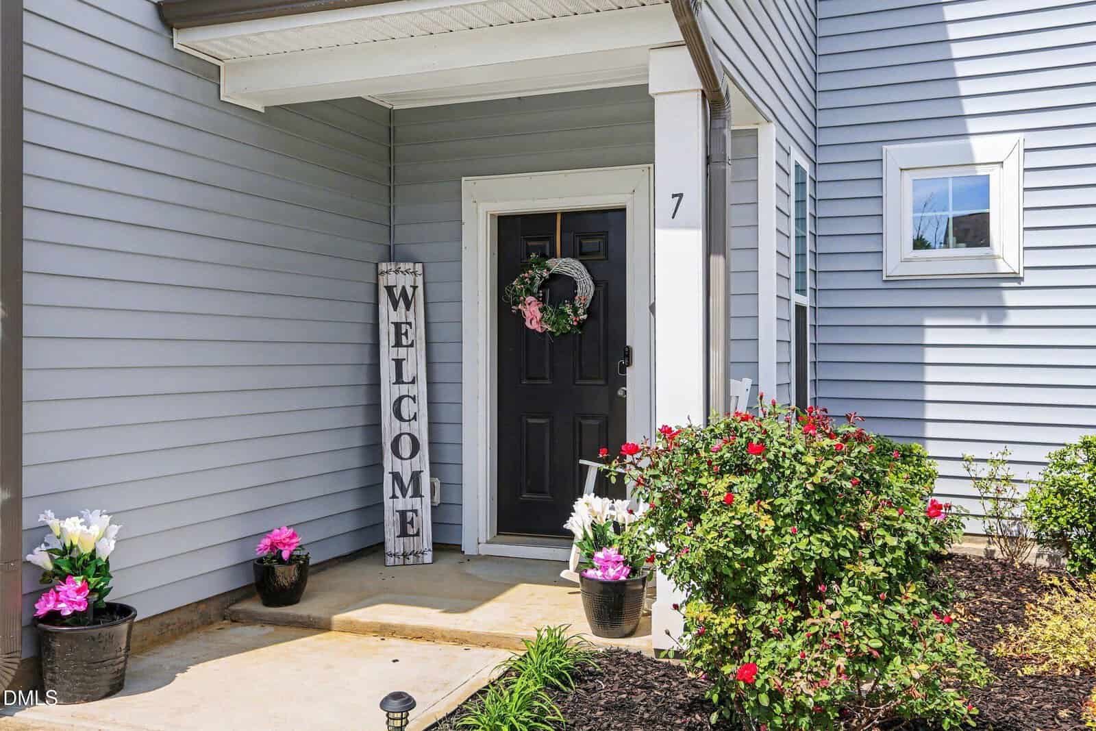 A front porch with a black door, a wreath with pink flowers, a large vertical "WELCOME" sign, and potted flowers; gray siding and blooming shrubs decorate the entryway.