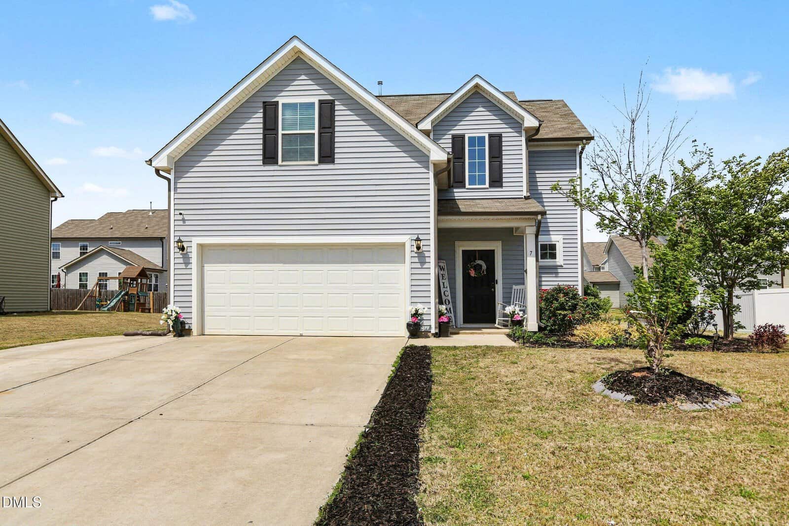 A two-story gray house with white trim at 7 Bromley Street, Franklinton features a double garage, wide concrete driveway, small front porch with potted plants, and a landscaped yard with a tree and mulch under a clear blue sky.