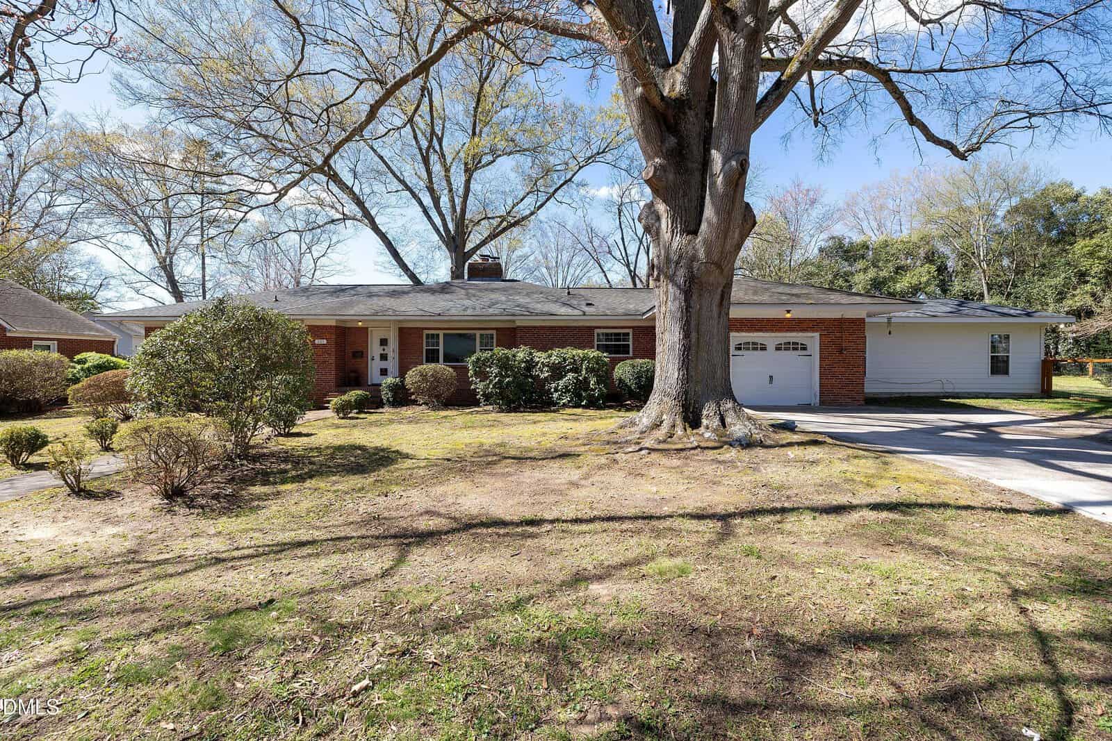 Single-story brick house with attached two-car garage, large front yard, and mature trees. Bushes line the front of the house, and concrete driveways are visible on both sides. Sky is clear with some clouds.