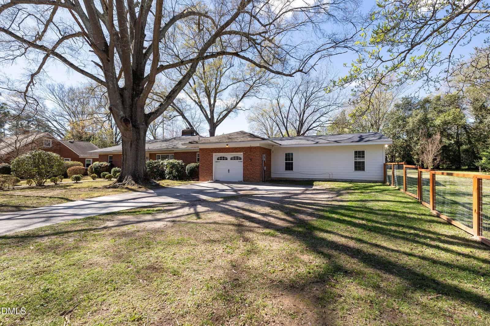 Single-story house with a brick and white exterior, two-car garage, large tree in the front yard, a paved driveway, and a fenced area on the right, set against a backdrop of trees and blue sky.