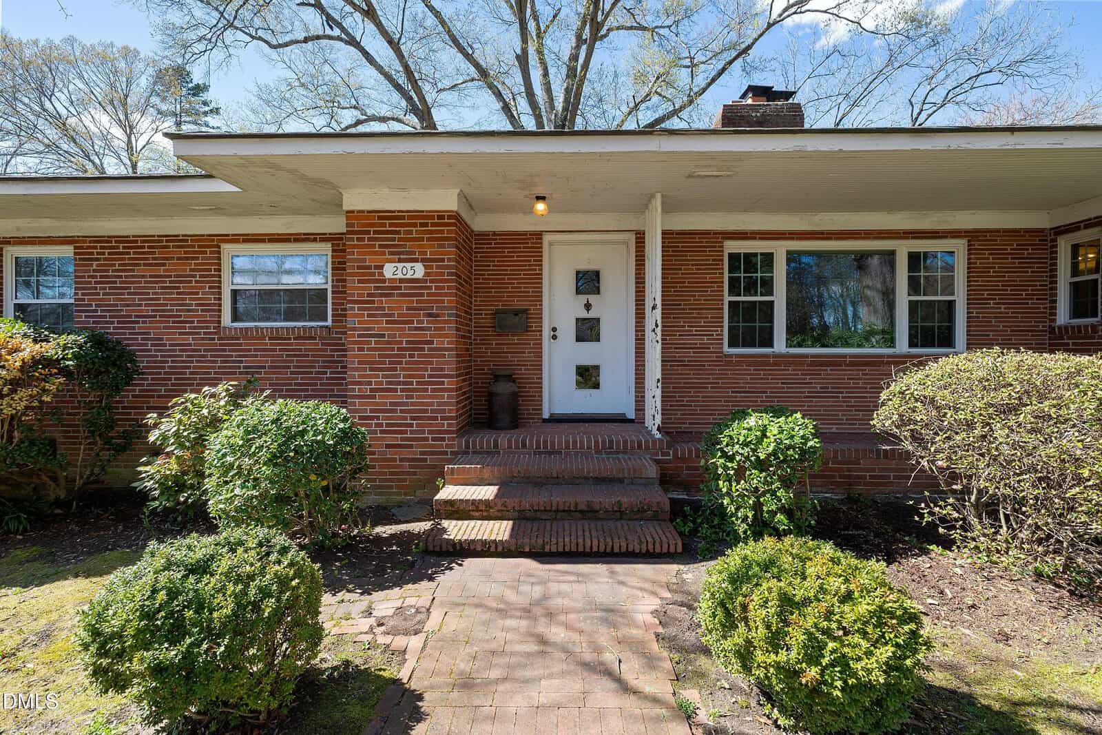 Single-story red brick house with a covered front porch, white door, two steps leading up to the entrance, two large windows, shrubs, and a small walkway in the front yard.