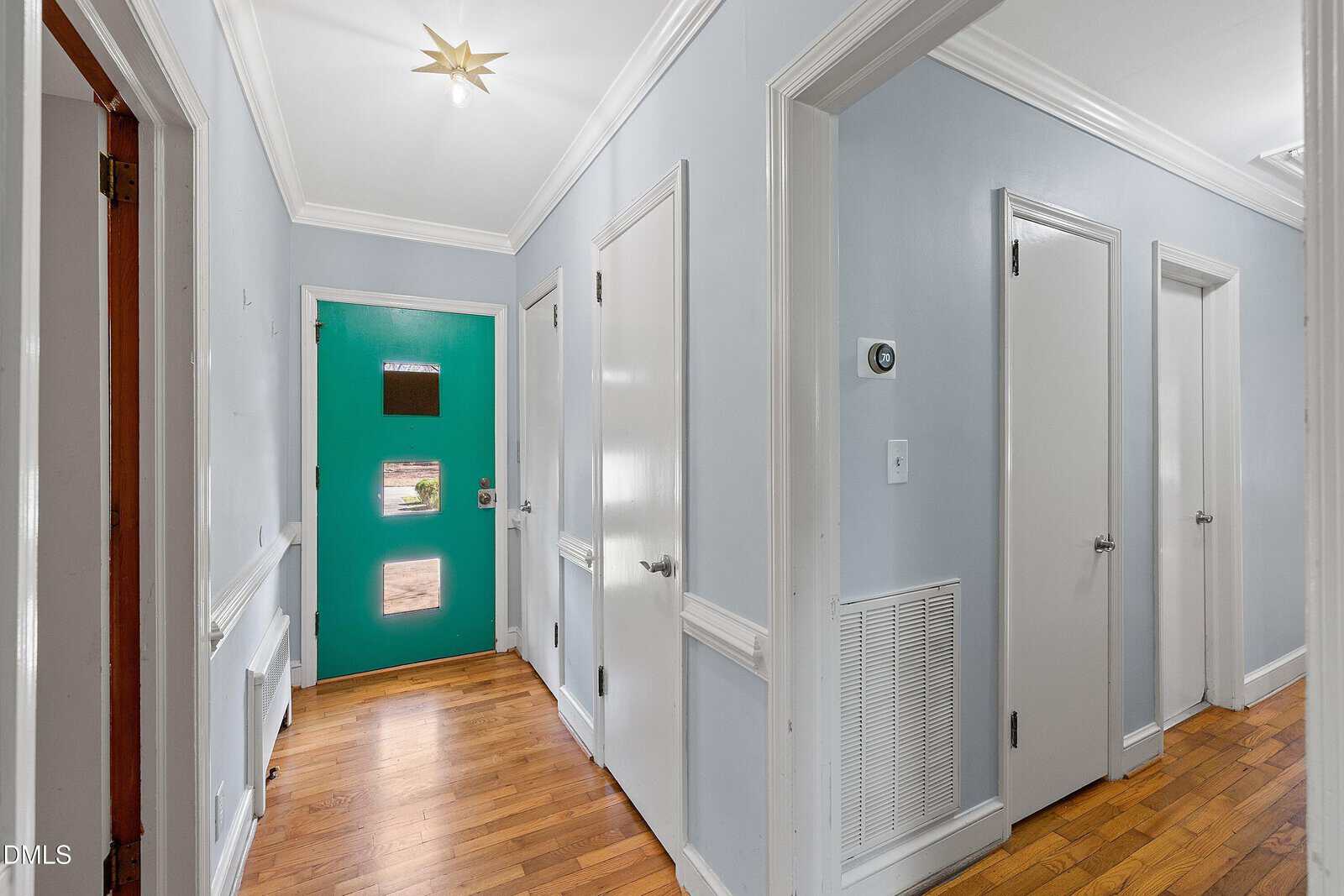 A hallway with hardwood floors, light blue walls, white trim, and three white doors. A turquoise front door with three small windows is at the end of the hall. A star-shaped ceiling light fixture is overhead.