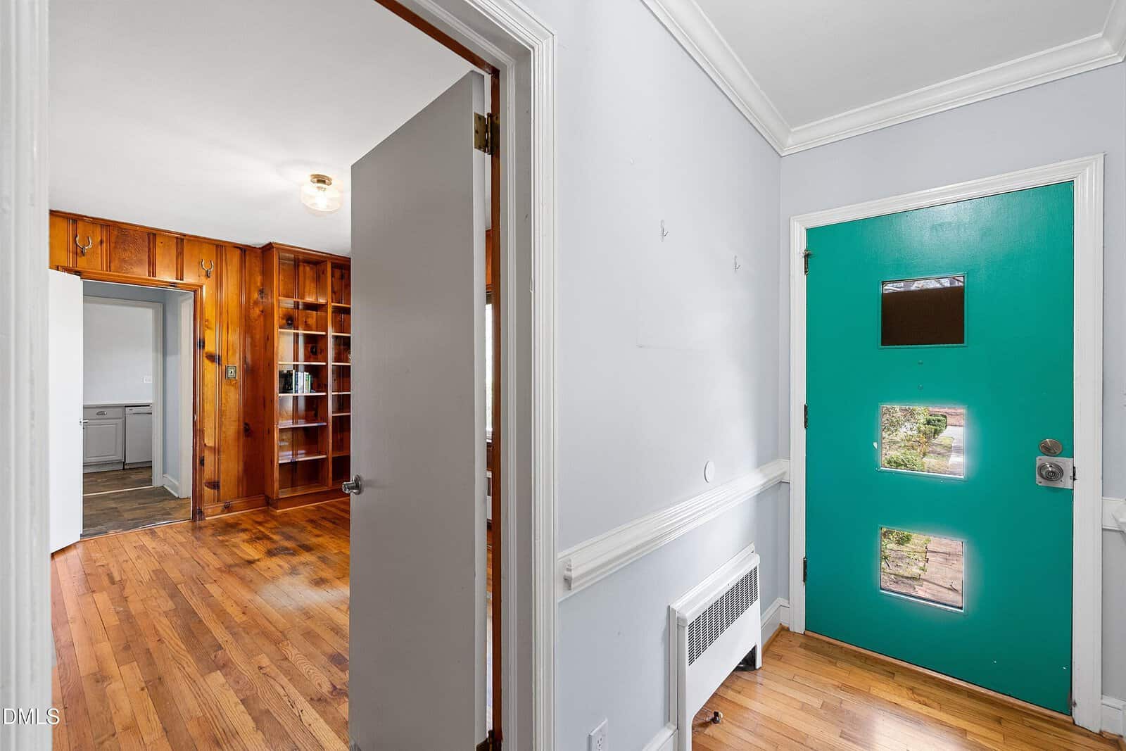 A turquoise front door with two square windows opens to a hallway with light gray walls, wood floors, and white trim. An adjacent room features wood-paneled walls and built-in shelves.