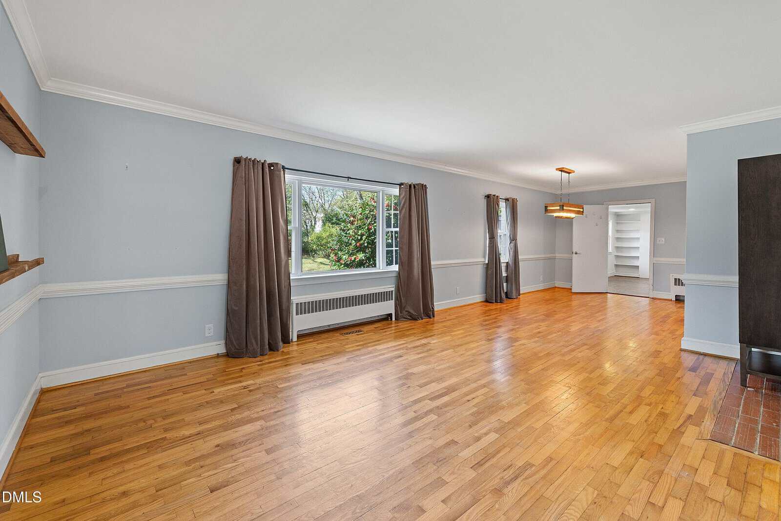 Spacious living and dining area with light gray walls, hardwood floors, large window with brown curtains, and modern light fixture. Natural light fills the room.