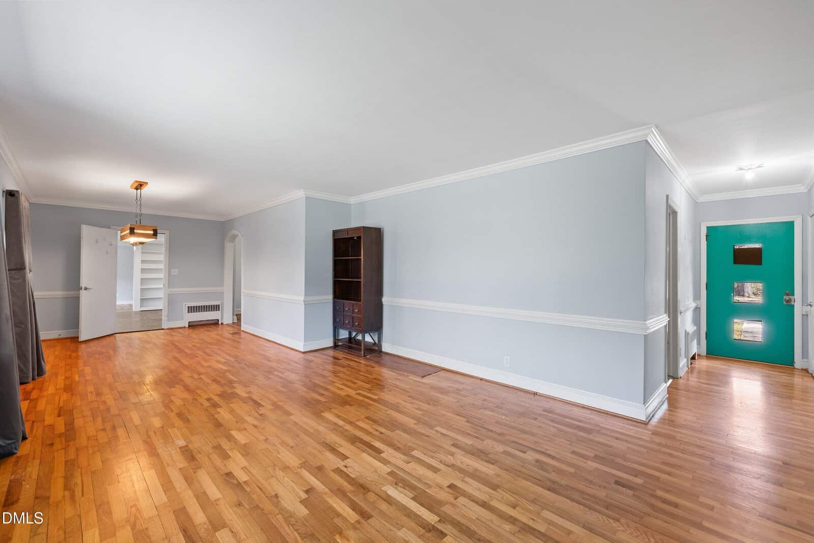 Spacious living room with hardwood floors, light gray walls, white trim, and crown molding. A bookcase stands against one wall. To the left, a dining area is visible; a teal front door is at the far end of a hallway on the right.