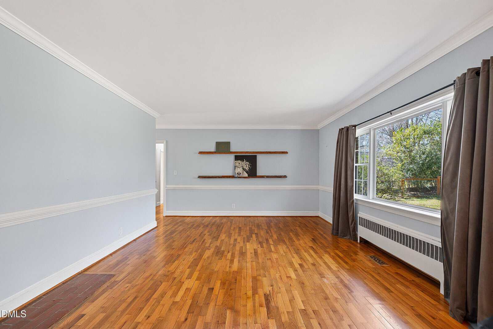 Empty living room with light gray walls, white trim, hardwood floors, three wooden shelves on the far wall, and a large window with brown curtains overlooking a green yard.
