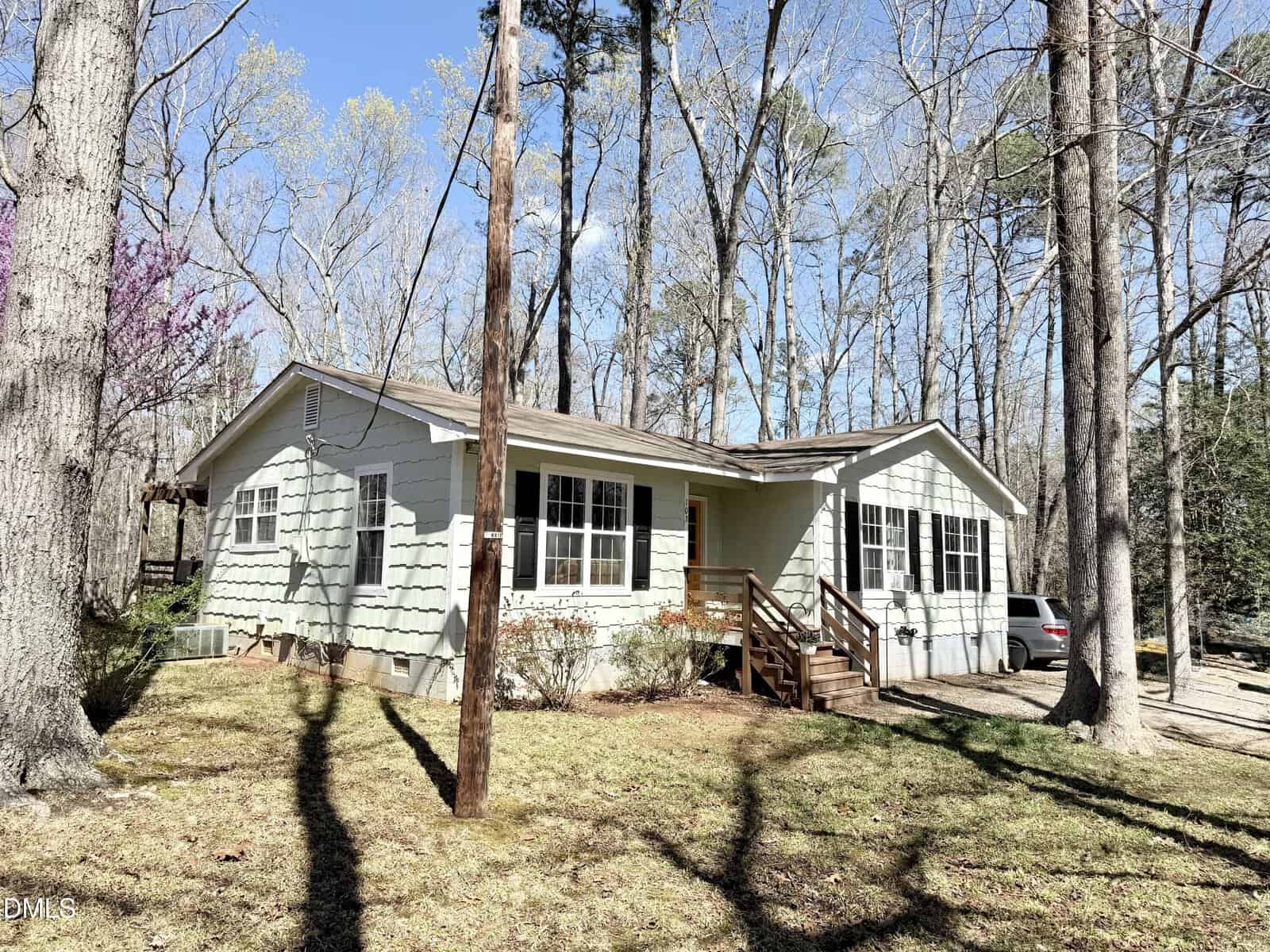 A single-story light green house with white trim, a small front porch with wooden steps, and multiple windows is surrounded by tall trees. A car is parked on the dirt driveway to the right. The yard is mostly shaded.