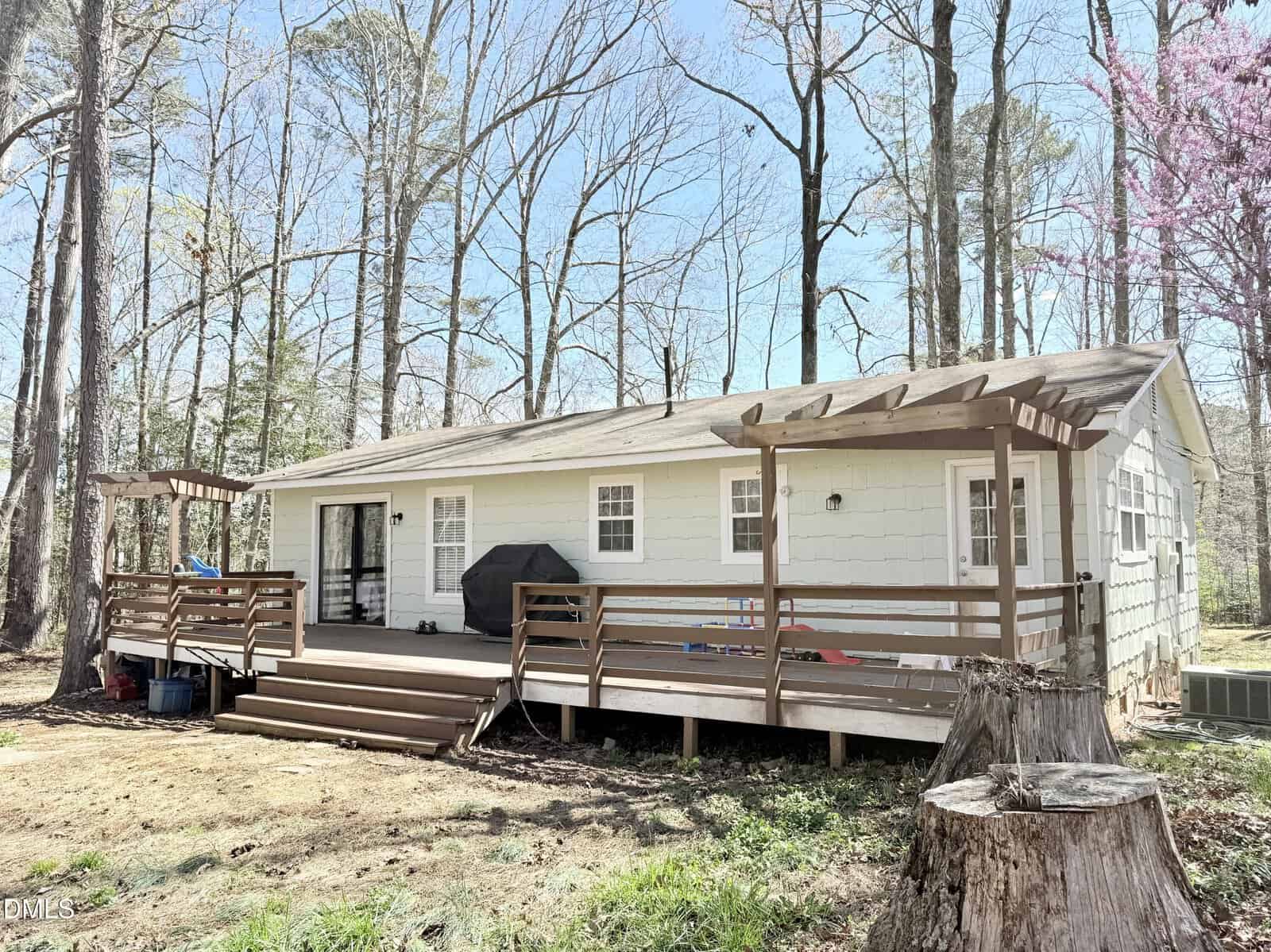 A single-story house with white siding and a wooden deck featuring two pergolas. The deck has railings, a grill, and children's toys. Leafless trees and a tree stump are visible in the yard on a sunny day.