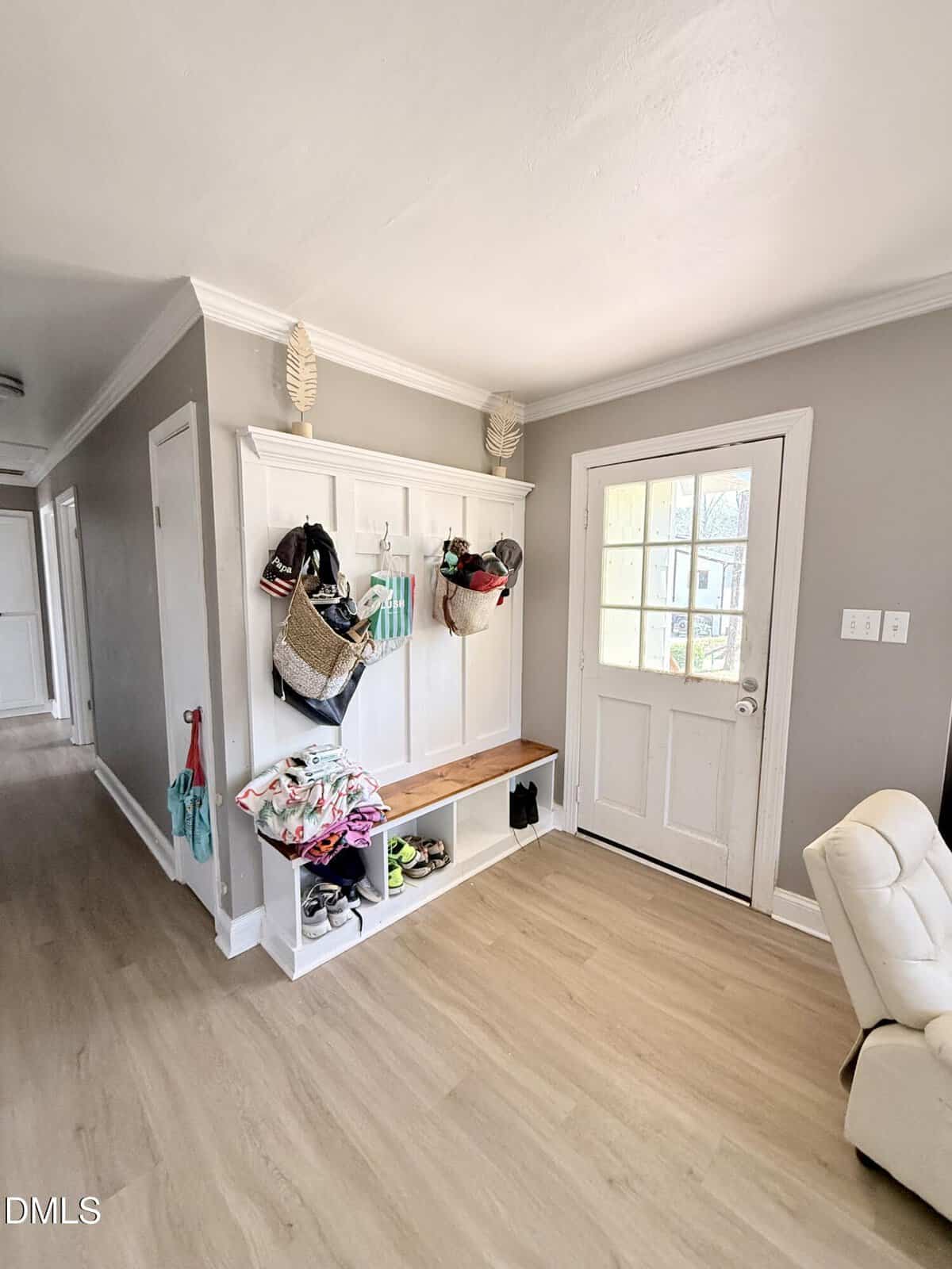 A hallway with light wood floors, gray walls, and white trim features a white storage bench with baskets, shoes, bags, and hats. A white door with glass panels is on the right, and a hallway extends to the left.
