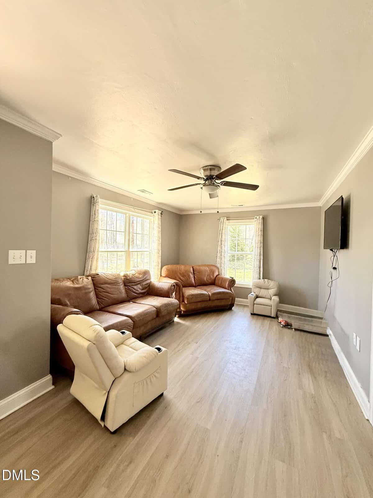 A living room with light wood flooring, gray walls, a ceiling fan, two brown leather sofas, a beige recliner, wall-mounted TV, and two windows with light curtains. Natural light fills the space.