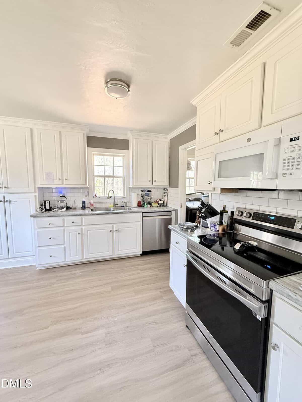 Modern kitchen with white cabinets, stainless steel appliances, a microwave above the stove, a dishwasher, and a window over the sink. Light wood flooring and neutral walls complete the bright space.