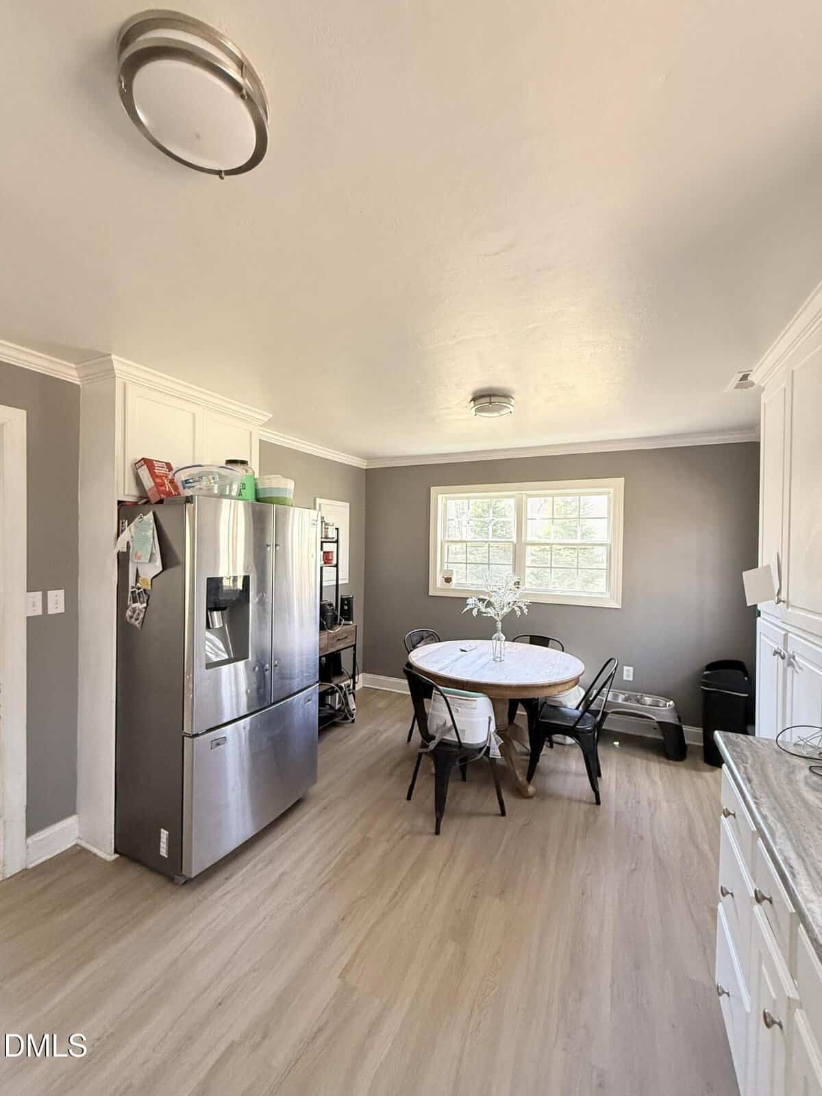 A kitchen with light wood floors, gray walls, white cabinets, a stainless steel refrigerator, a round table with four chairs, a vase with flowers, and two ceiling lights. Sunlight shines through a window above the table.