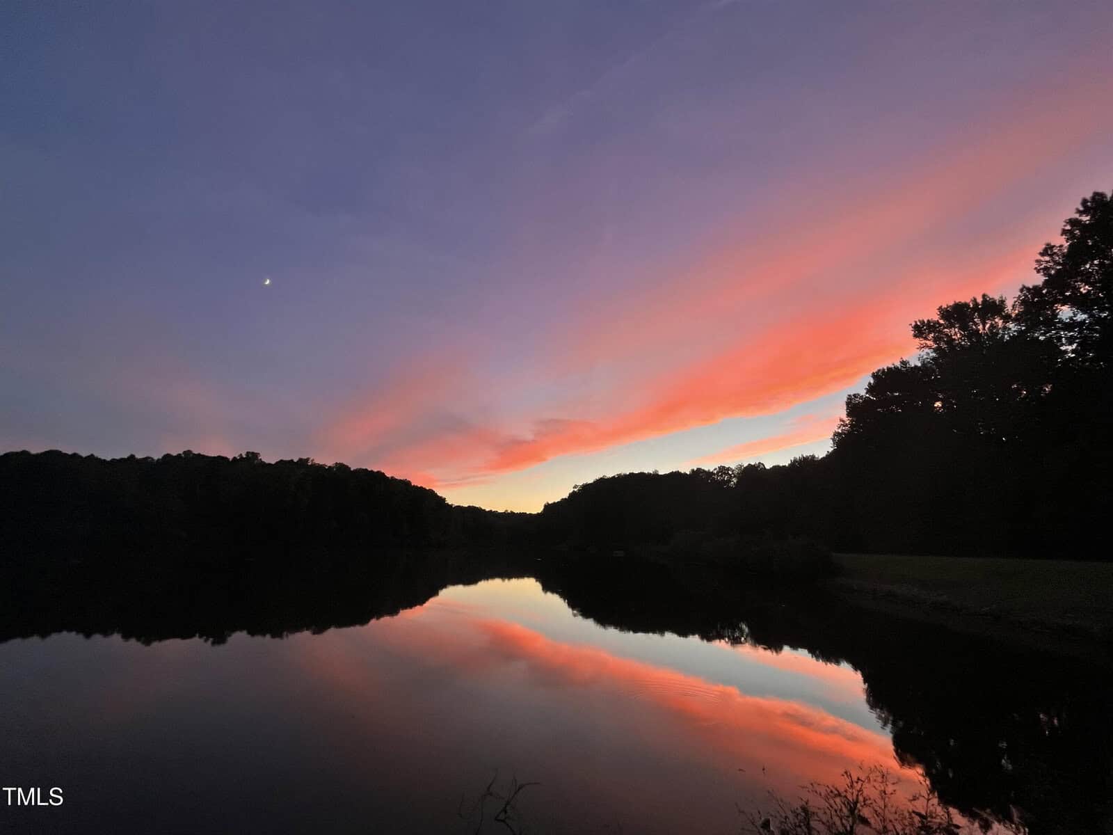 A serene lake at dusk reflects a colorful sky with pink, orange, and purple clouds. Silhouetted trees frame the horizon, and a small crescent moon is visible in the upper left corner.