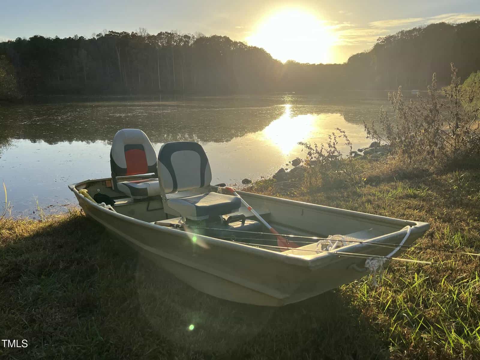 A small fishing boat with two seats is pulled up on the grassy shore of a calm lake at sunset, with trees and sunlight reflected on the water in the background.
