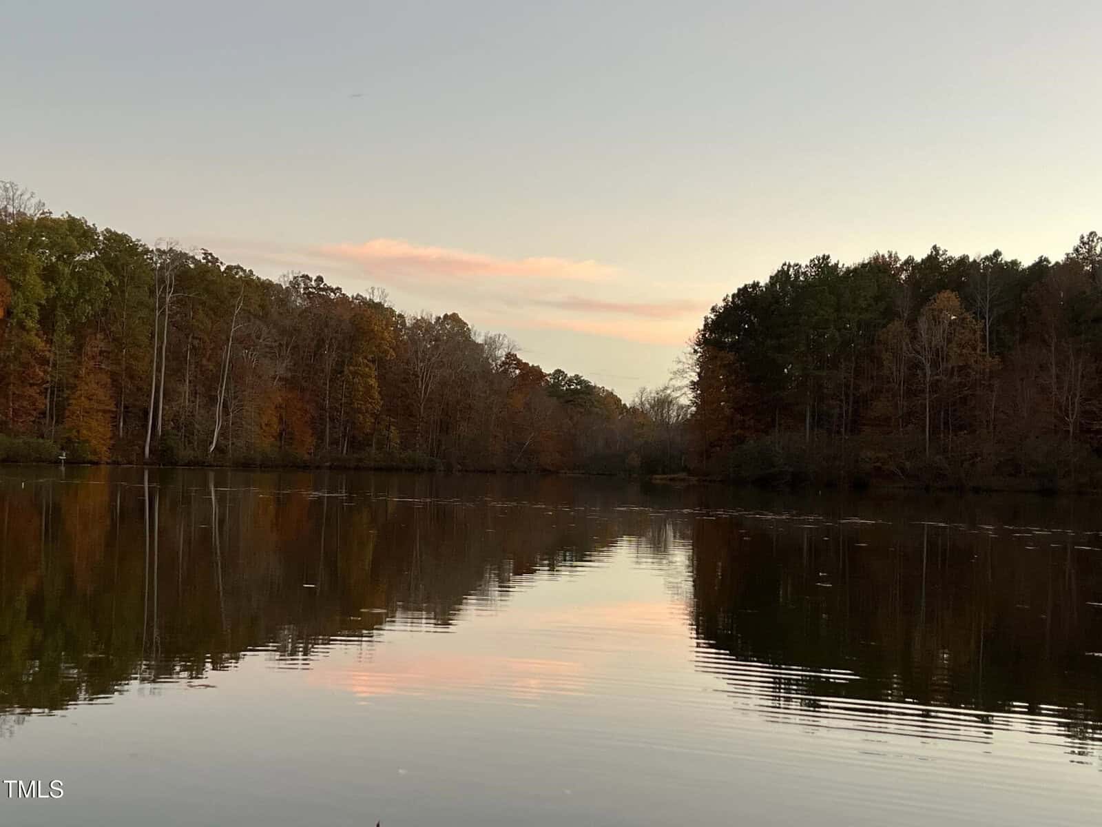 A calm lake reflects autumn trees with orange and green leaves under a pale evening sky with pink clouds. The shoreline is lined with dense forest.
