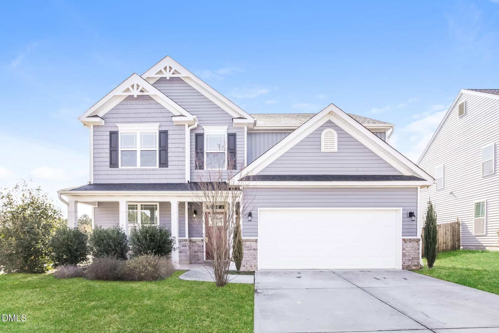 A two-story suburban house with gray siding, white trim, and a double garage. The front yard has green grass, bushes, and a small leafless tree. The sky is clear and blue.