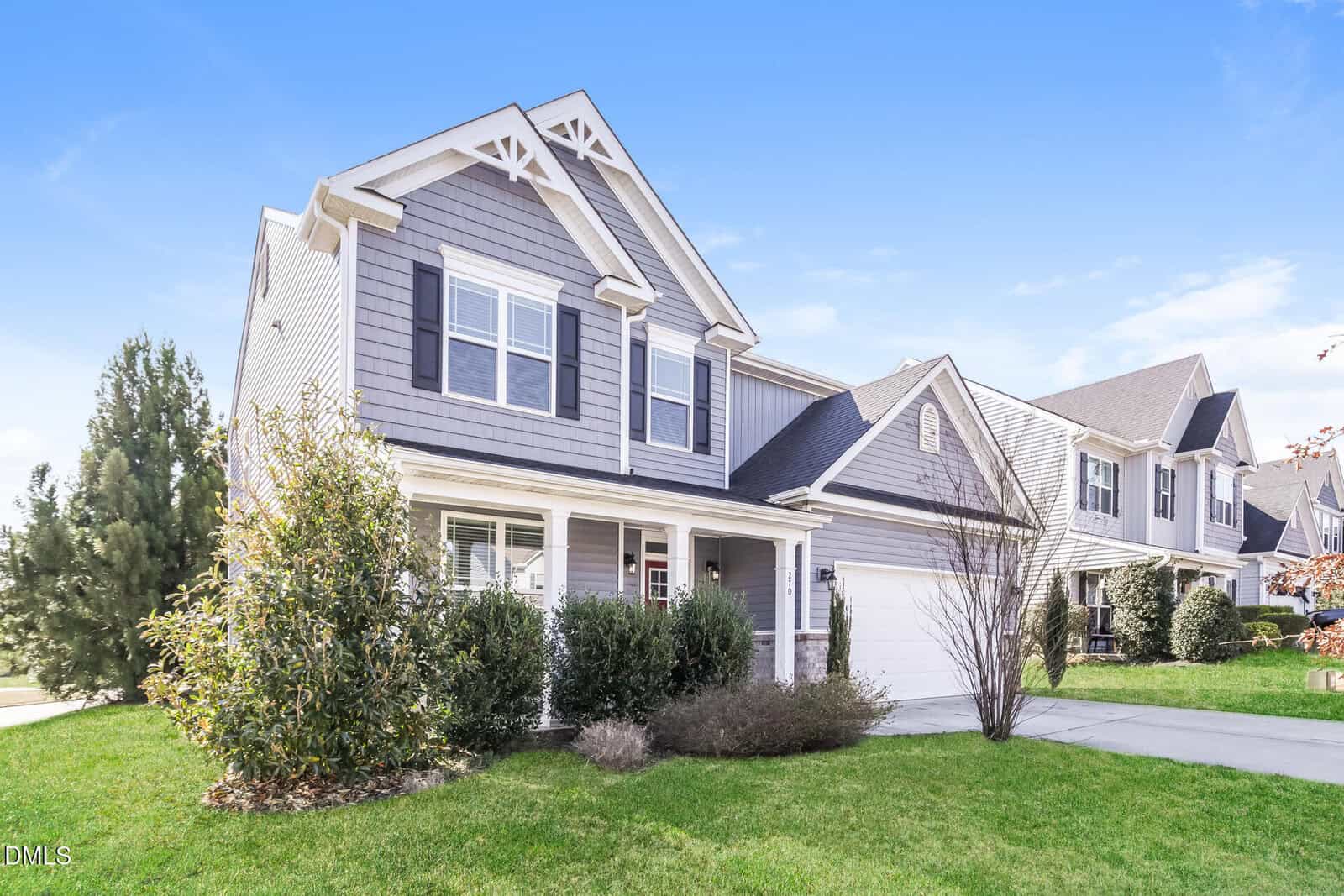 A two-story suburban house with gray siding, white trim, and a covered front porch. The home has an attached two-car garage, a concrete driveway, and neatly maintained shrubs and lawn. Other similar houses are nearby.
